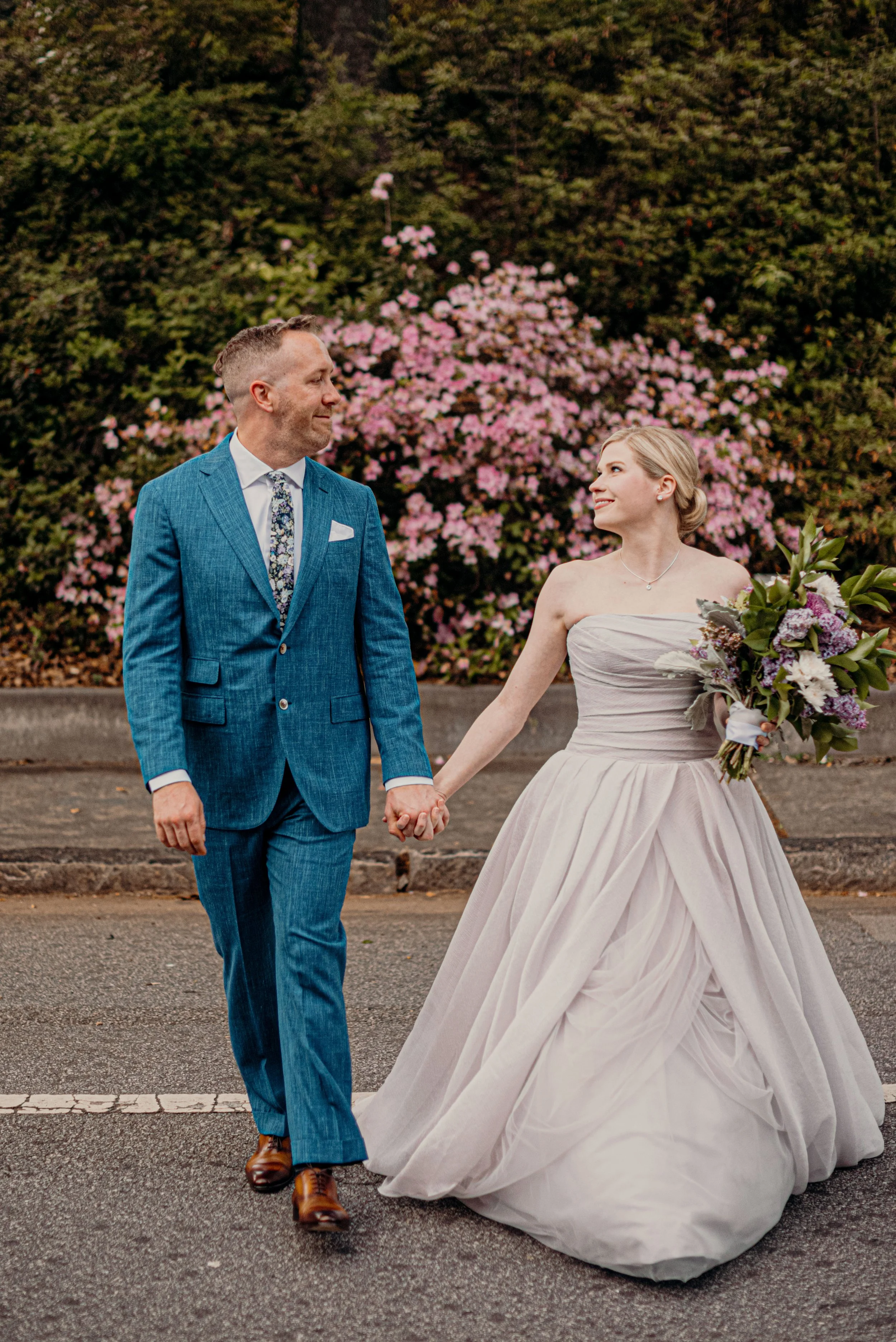 A bride and groom holding hands and walking outside during a wedding. The groom is in a blue suit and the bride is in a strapless white wedding dress holding a bouquet of flowers. Pink flowering bushes are in the background.