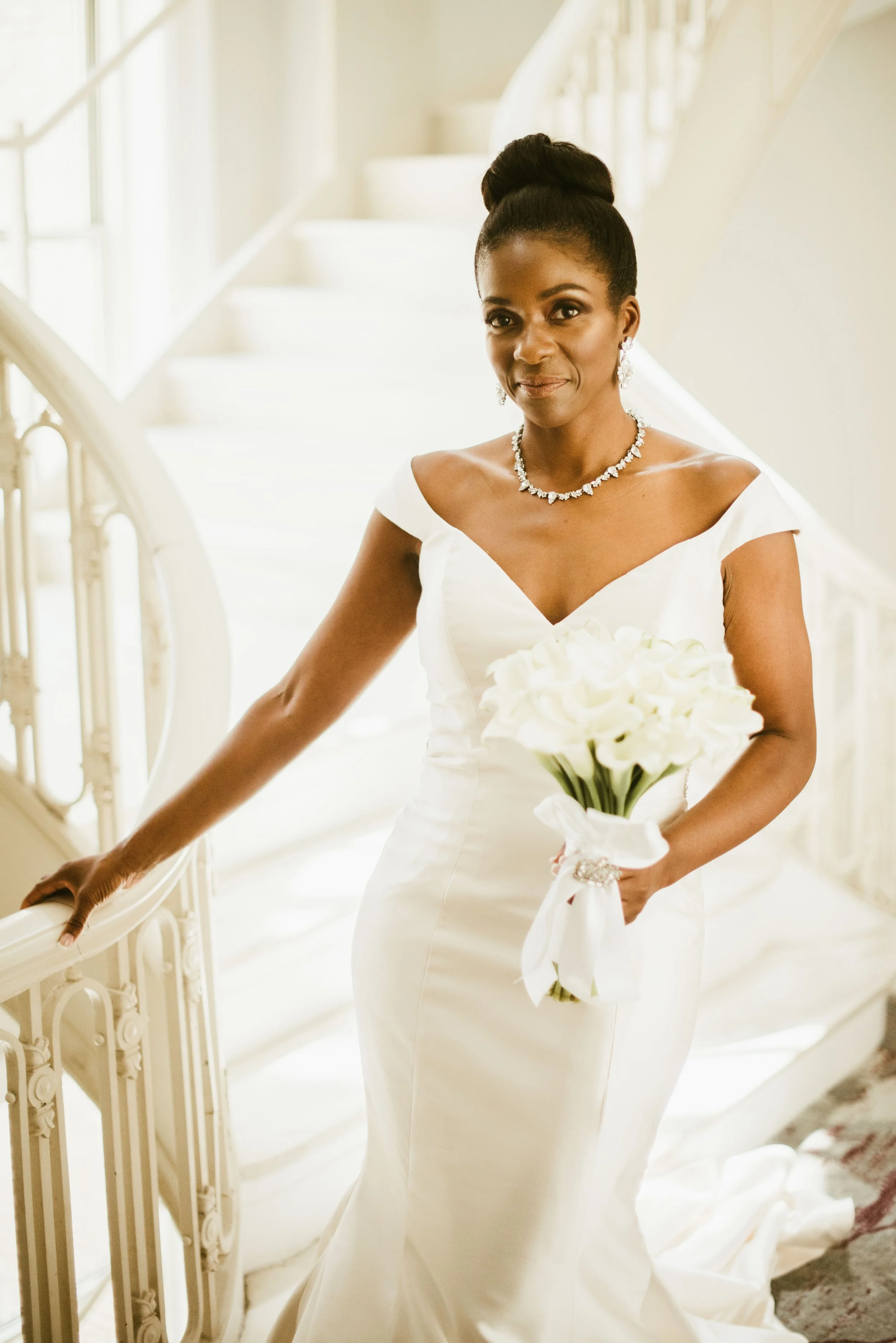 A woman in a white wedding dress holding a bouquet of white flowers indoors, standing next to a staircase with ornate railing.