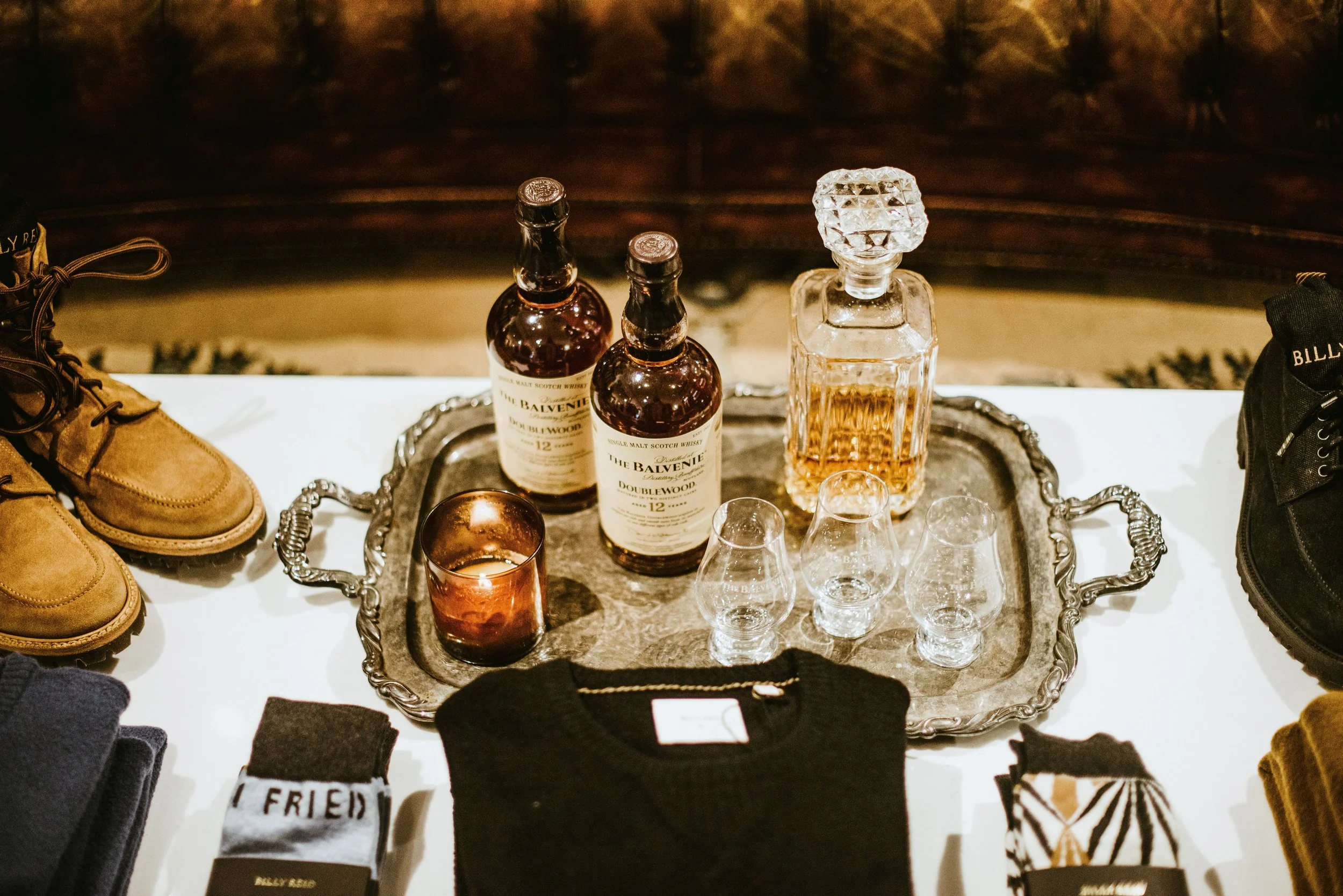 A display table with perfume bottles, a candle, glasses, and clothing items, including boots on both sides, on a white surface with a dark background.