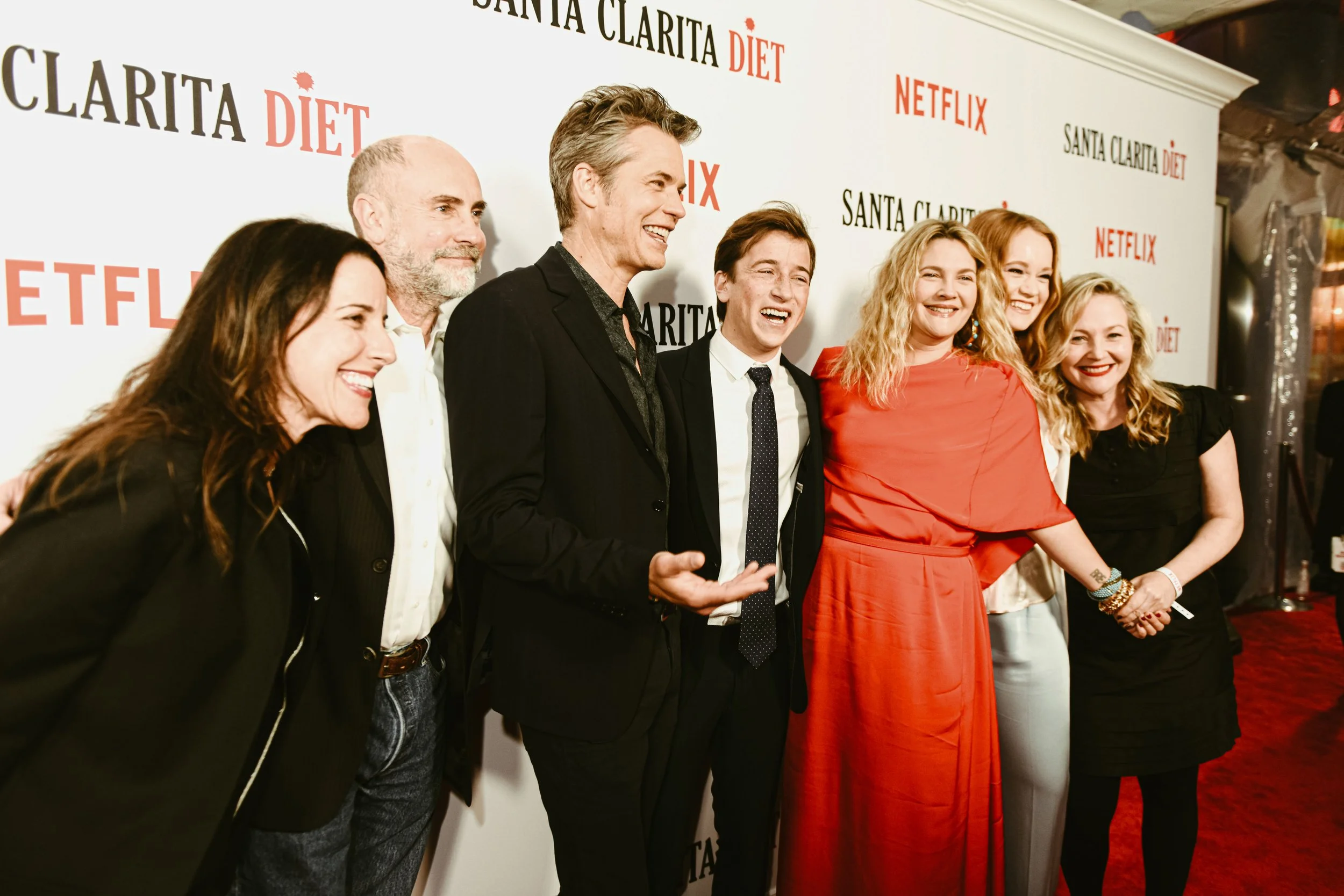 Group of people at a red carpet event standing in front of a white backdrop with 'Santa Clarita Diet' and 'Netflix' logos, smiling and posing for photos.