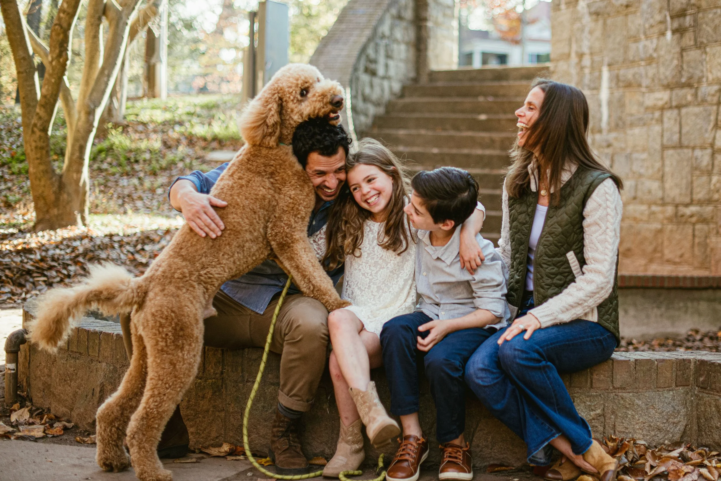 A family of five, including two children, a man, a woman, and a large curly-haired dog, enjoying a hug and laughter outdoors near stone steps and a brick wall in autumn.