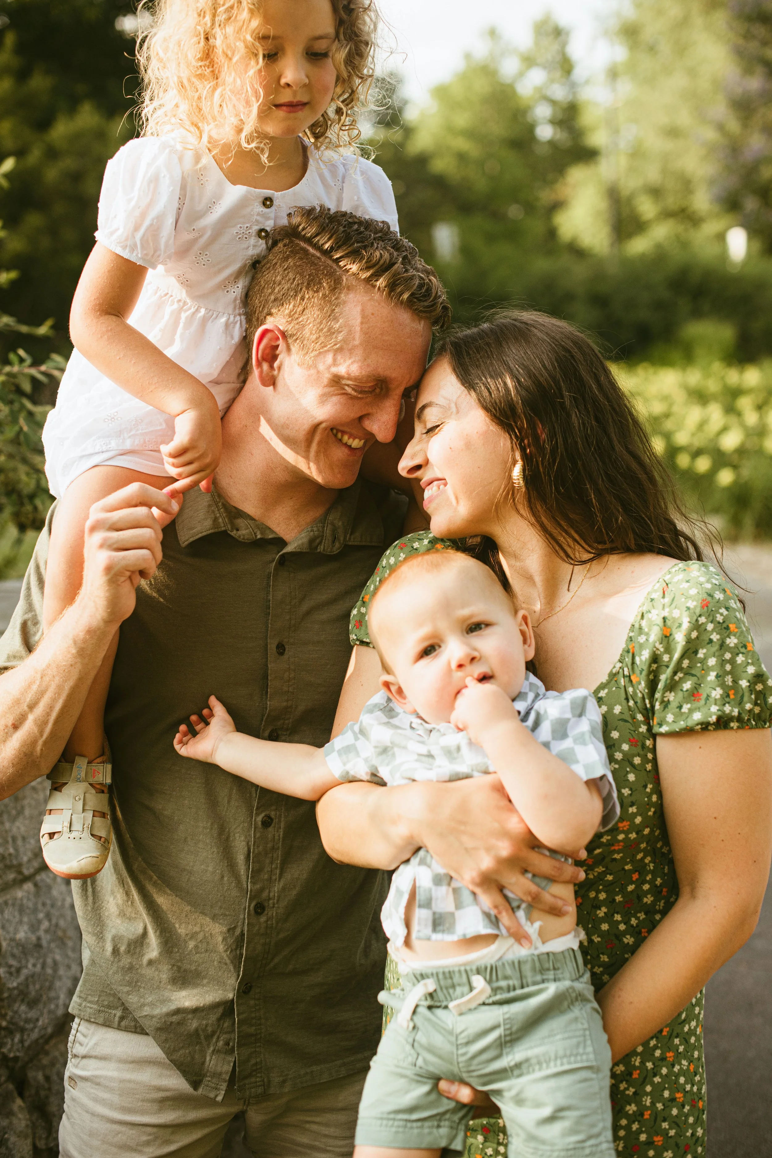 A happy family of four outdoors, with the father and mother smiling and embracing, the father holding the young son, and the daughter sitting on the father's neck.
