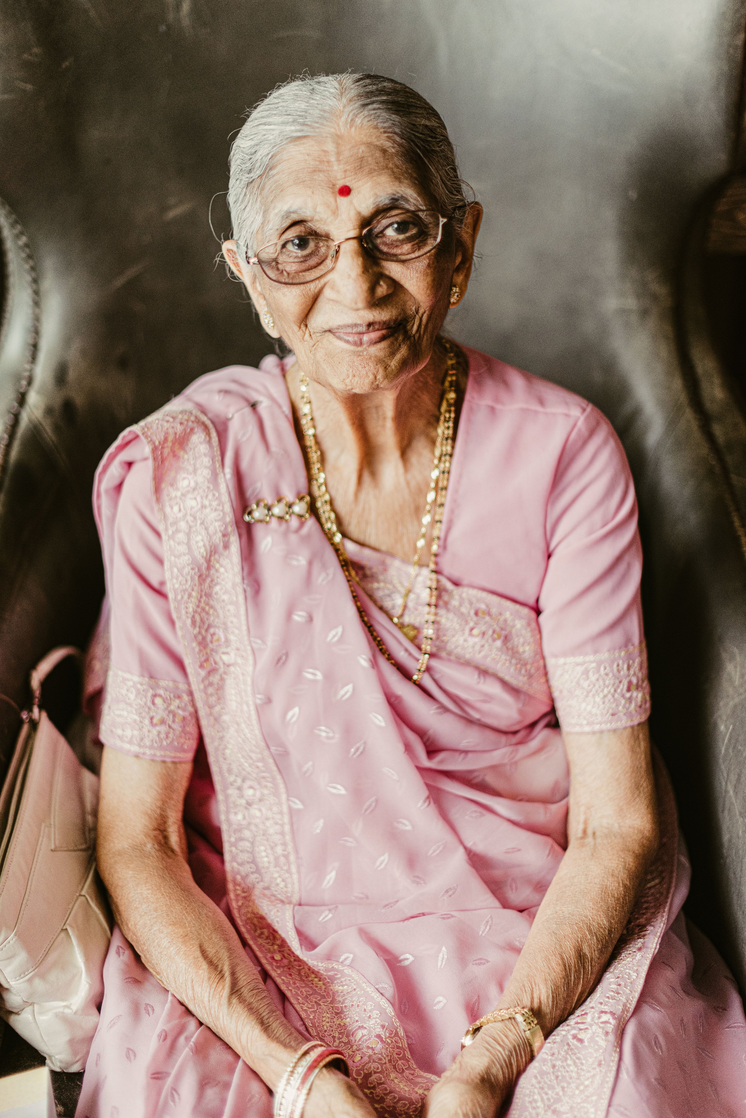 An elderly woman dressed in a pink traditional Indian sari with intricate embroidery, wearing gold jewelry, glasses, and a red bindi on her forehead, sitting on a chair with a neutral-colored background.
