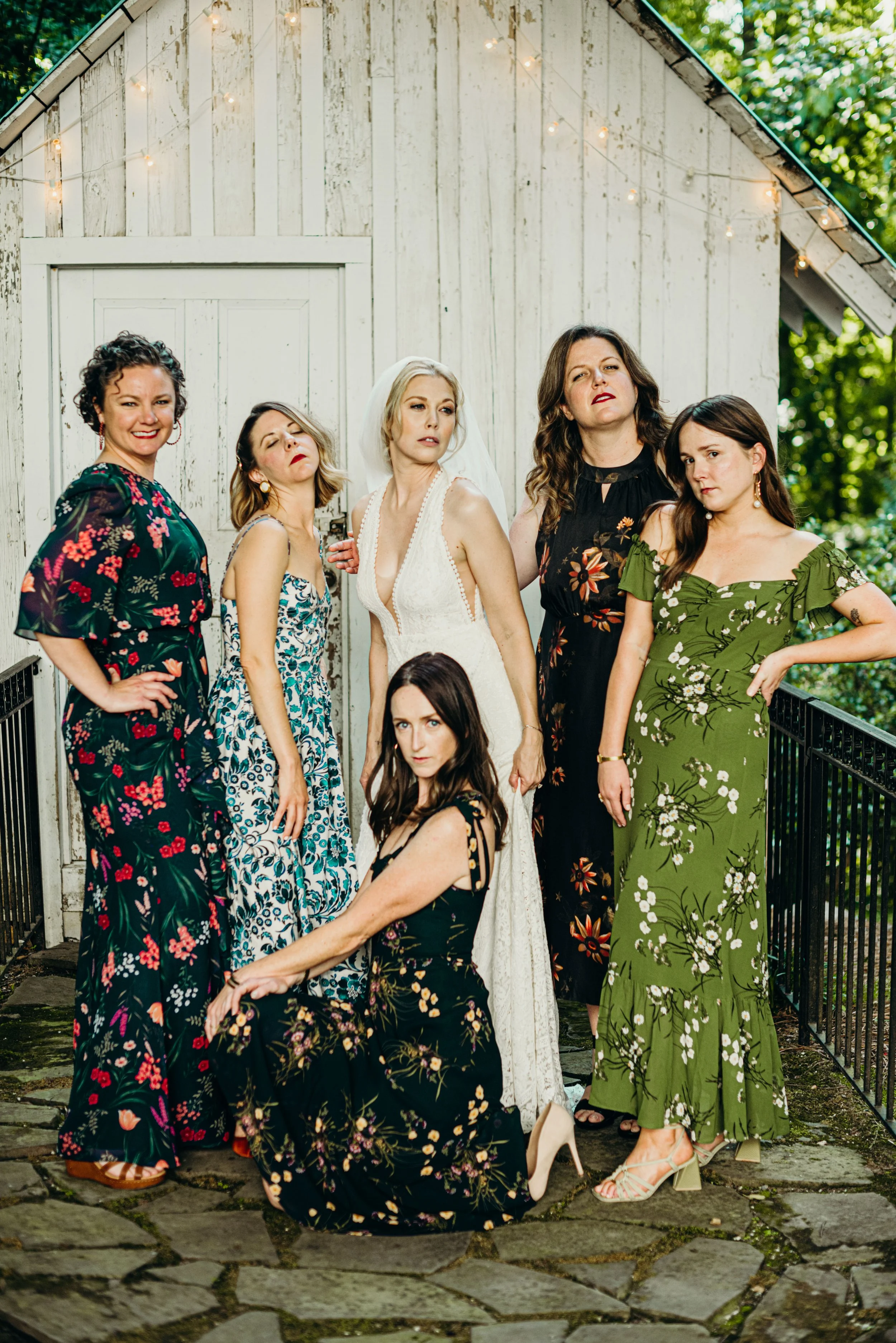 Group of six women standing outdoors in front of a rustic white wooden shed with string lights, casual poses, wearing floral dresses, some with heels, on a stone pathway with greenery in the background.