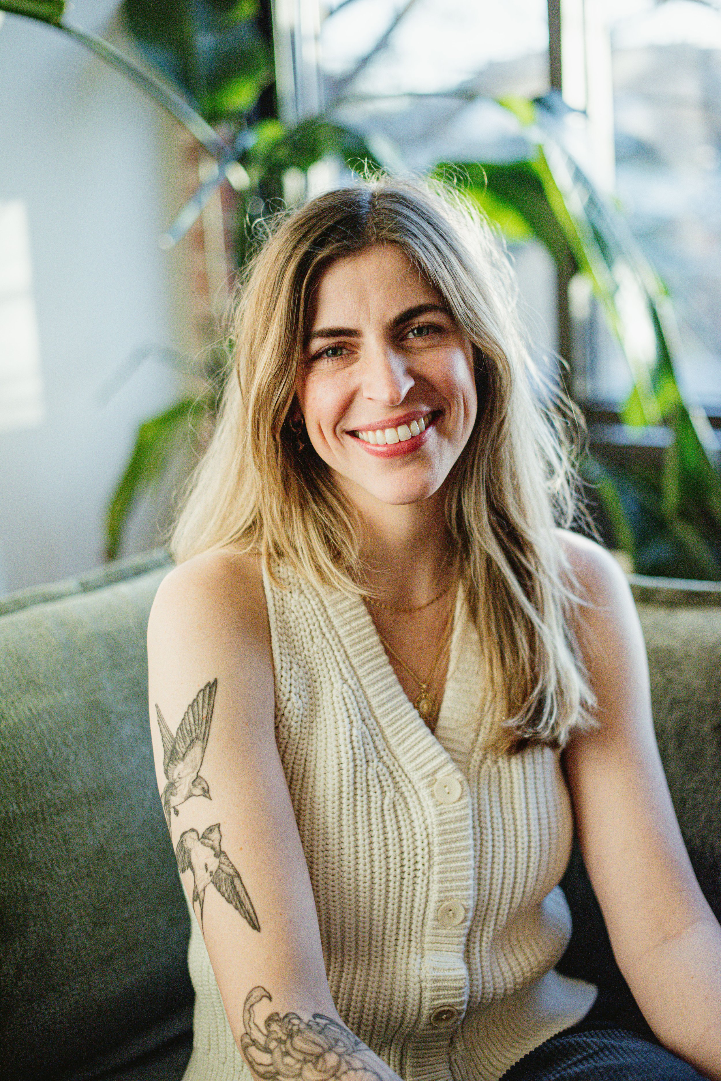 A young woman with long blonde hair smiling, sitting on a green sofa in a room with large windows, wearing a sleeveless cream-colored knitted top and displaying tattoos on her left arm.