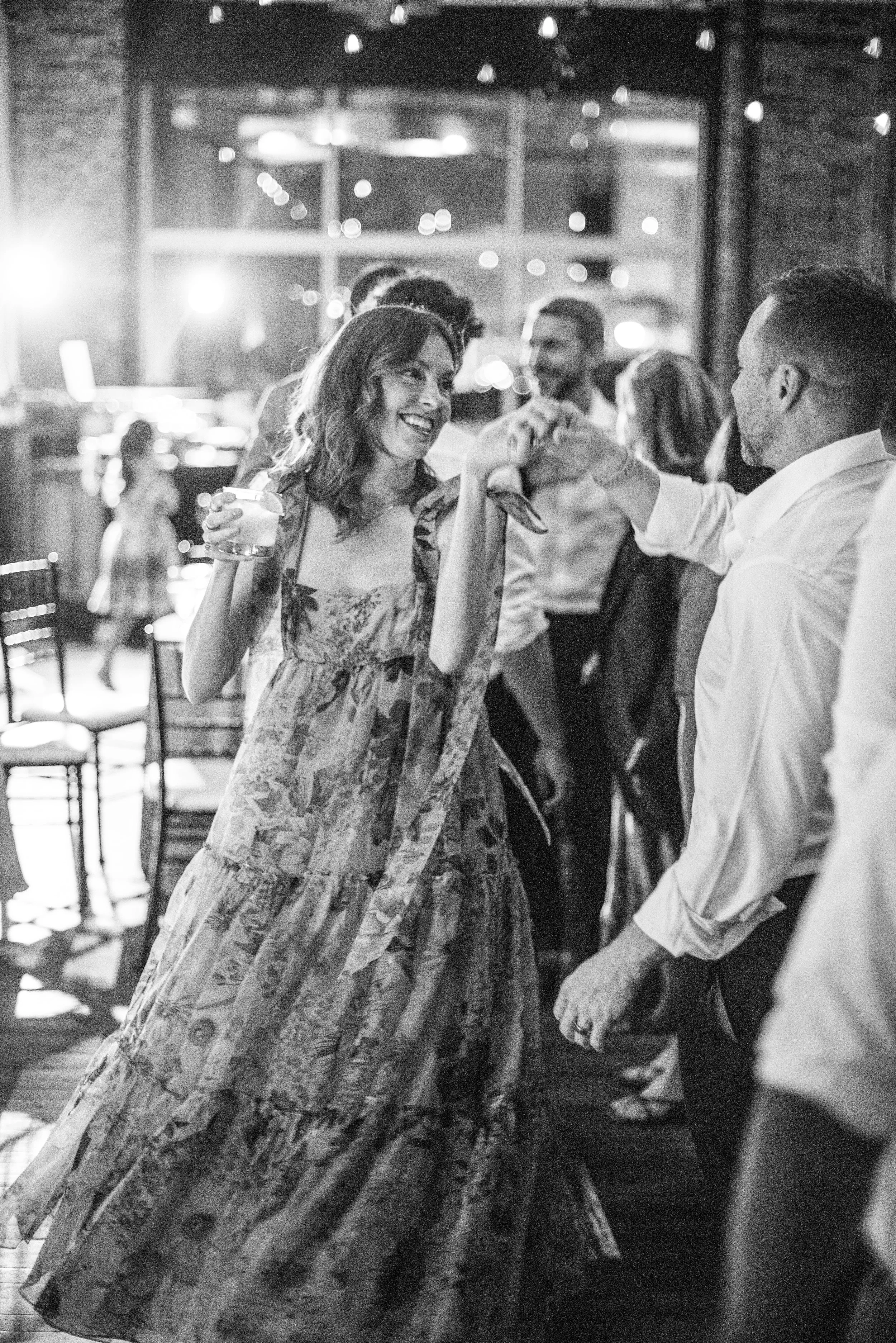 A woman in a floral dress holding a drink, smiling, and looking at a man in a white shirt at a social gathering or party held indoors.
