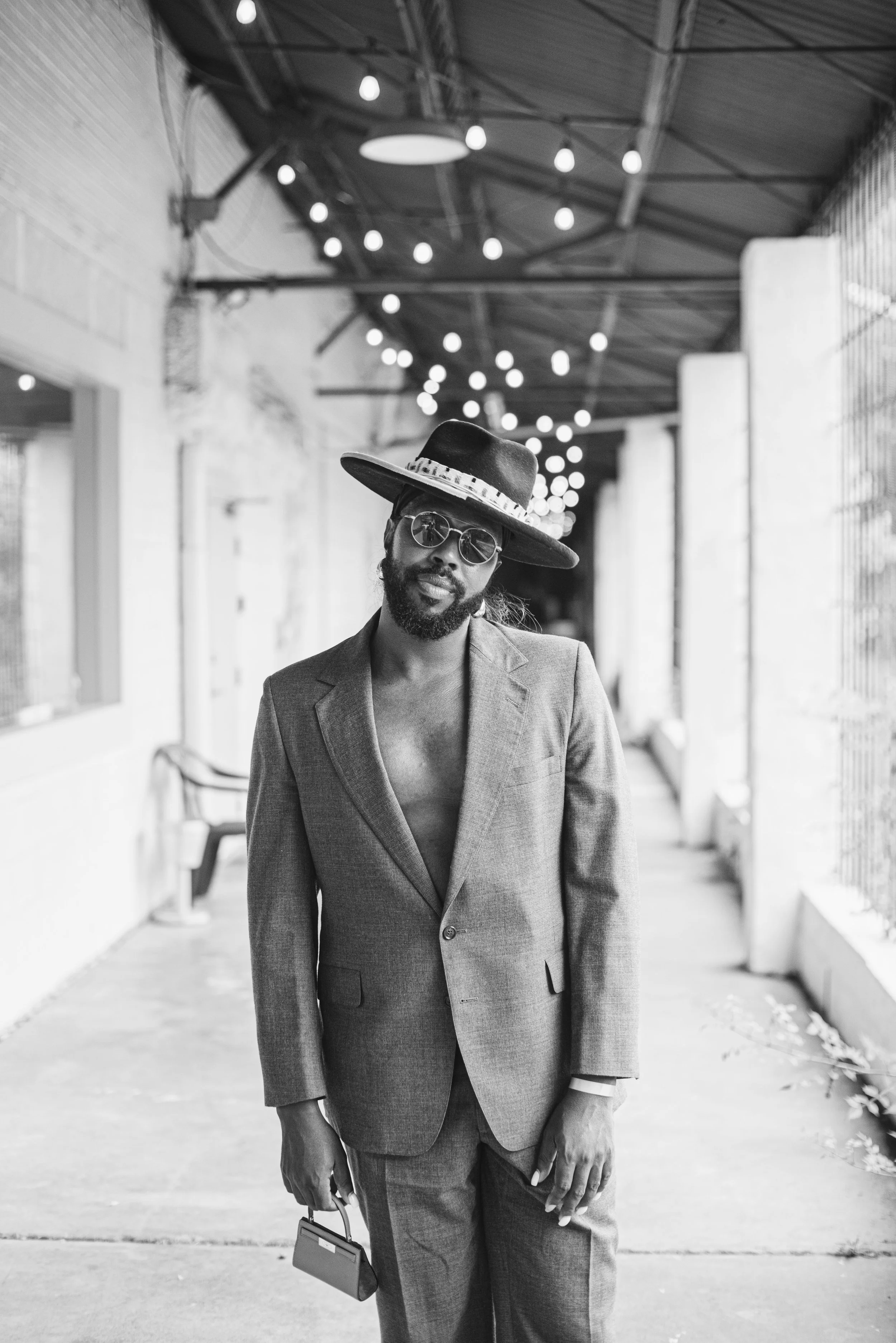 A man in a suit, wearing sunglasses, a wide-brimmed hat, and holding a small briefcase, standing outdoors in an urban environment with string lights hanging overhead.