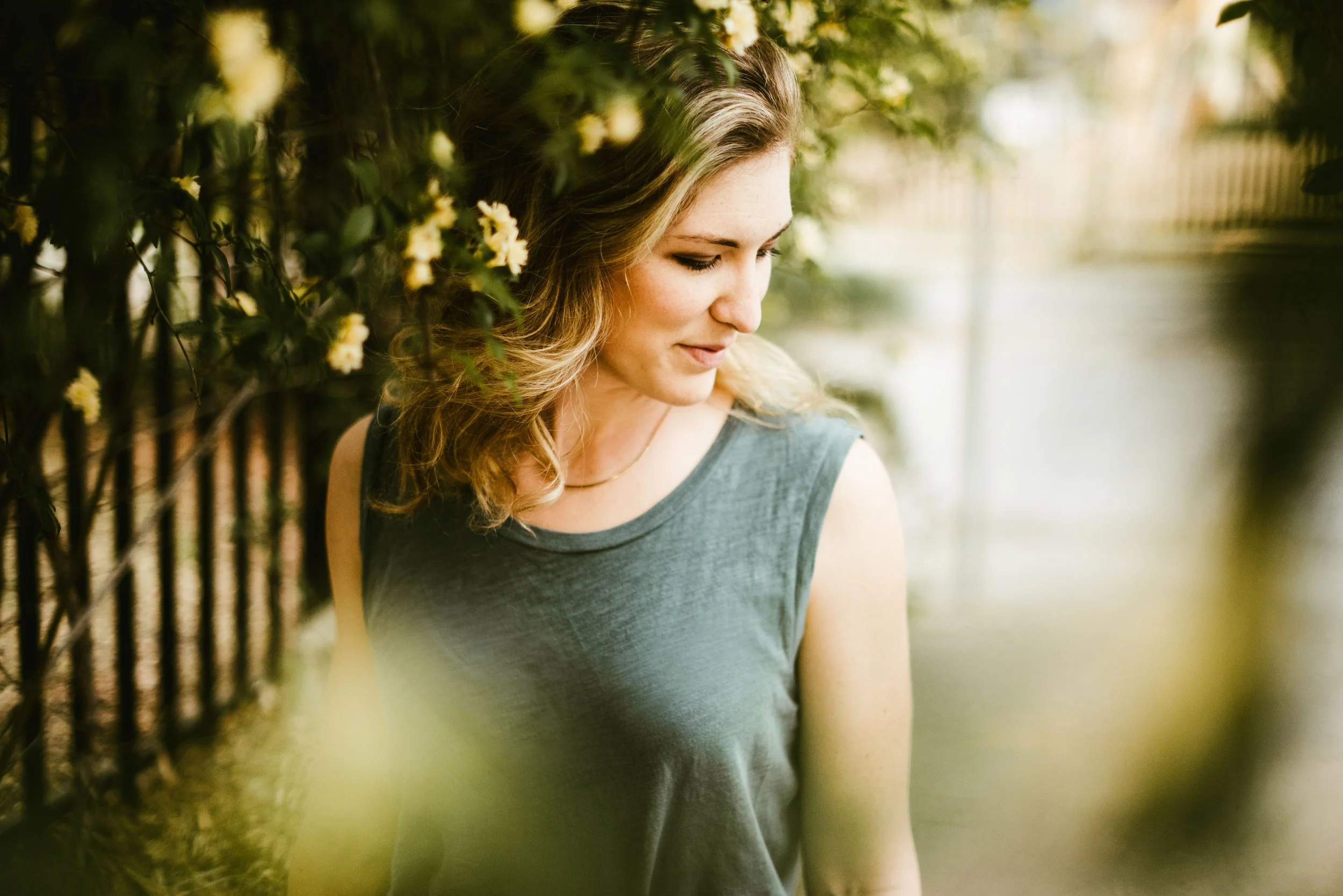 A woman with wavy light brown hair and a sleeveless top, standing outdoors near a fence covered with green foliage and small yellow flowers, with a blurred background.