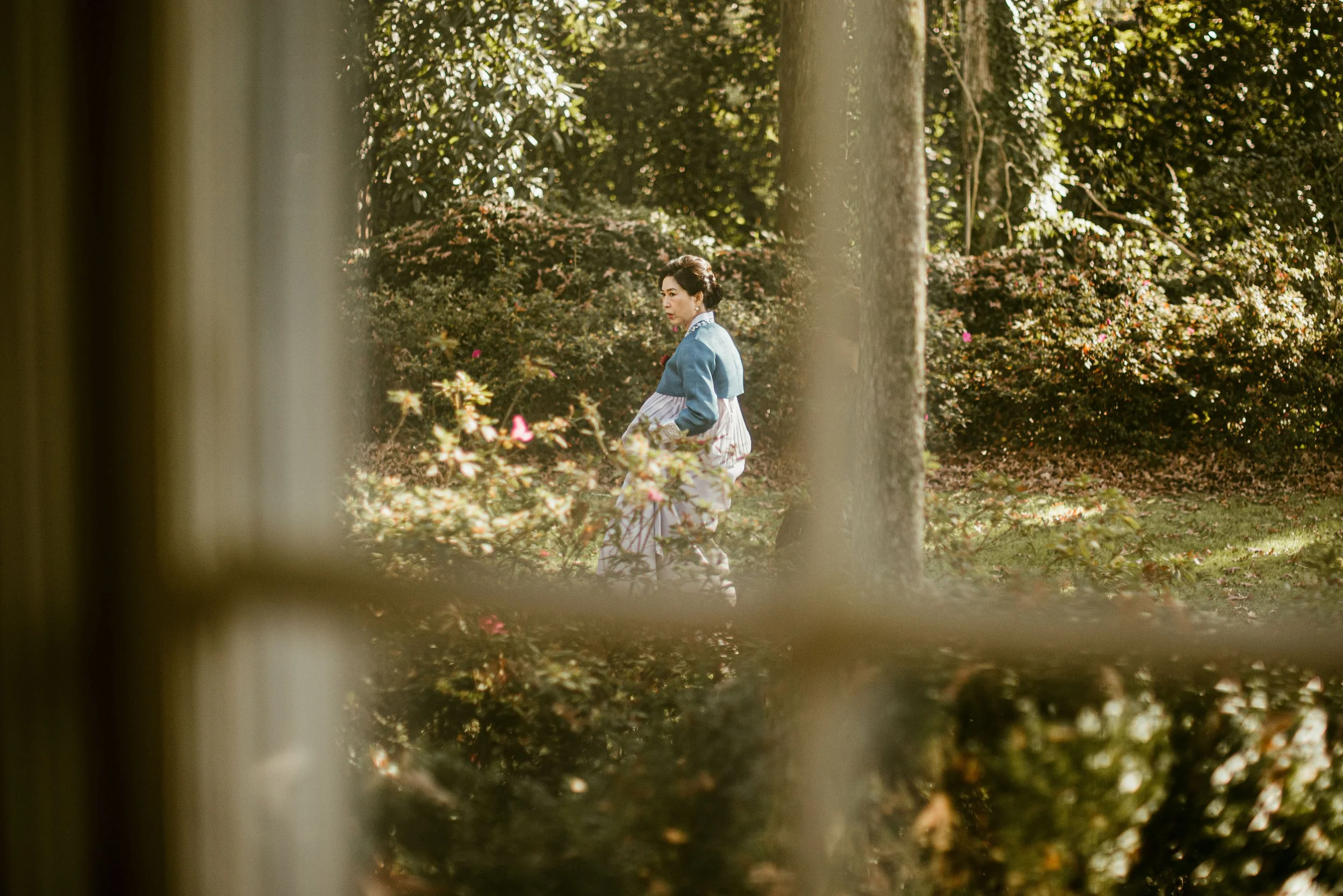 A woman in traditional kimono walking in a park with lush green trees and flowers, seen through a wooden fence.