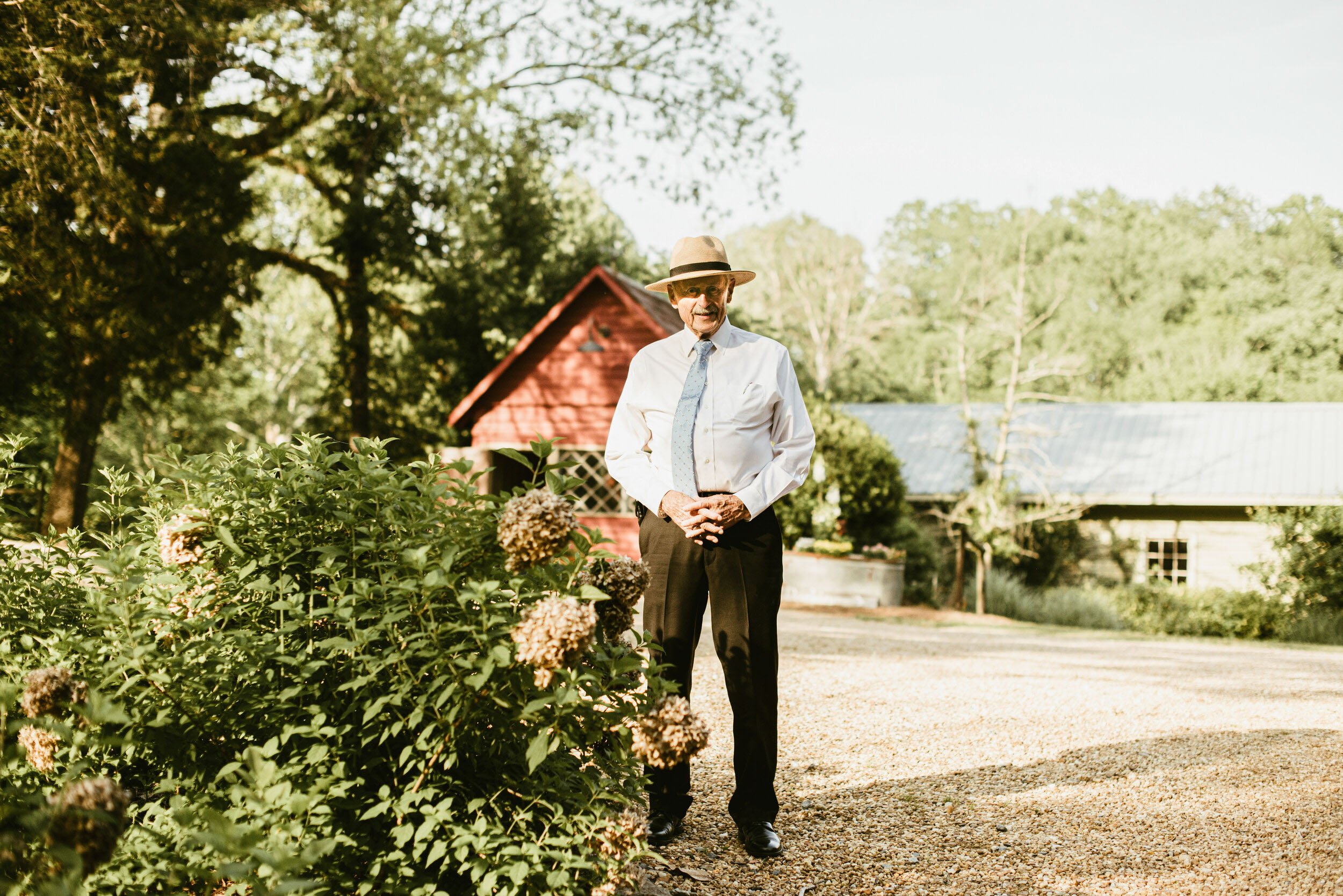 An elderly man wearing a white shirt, loosened tie, and a straw hat is standing outdoors on a gravel path near a garden with hydrangea bushes. There is a red wooden building with a porch and a house with a metal roof in the background, surrounded by 