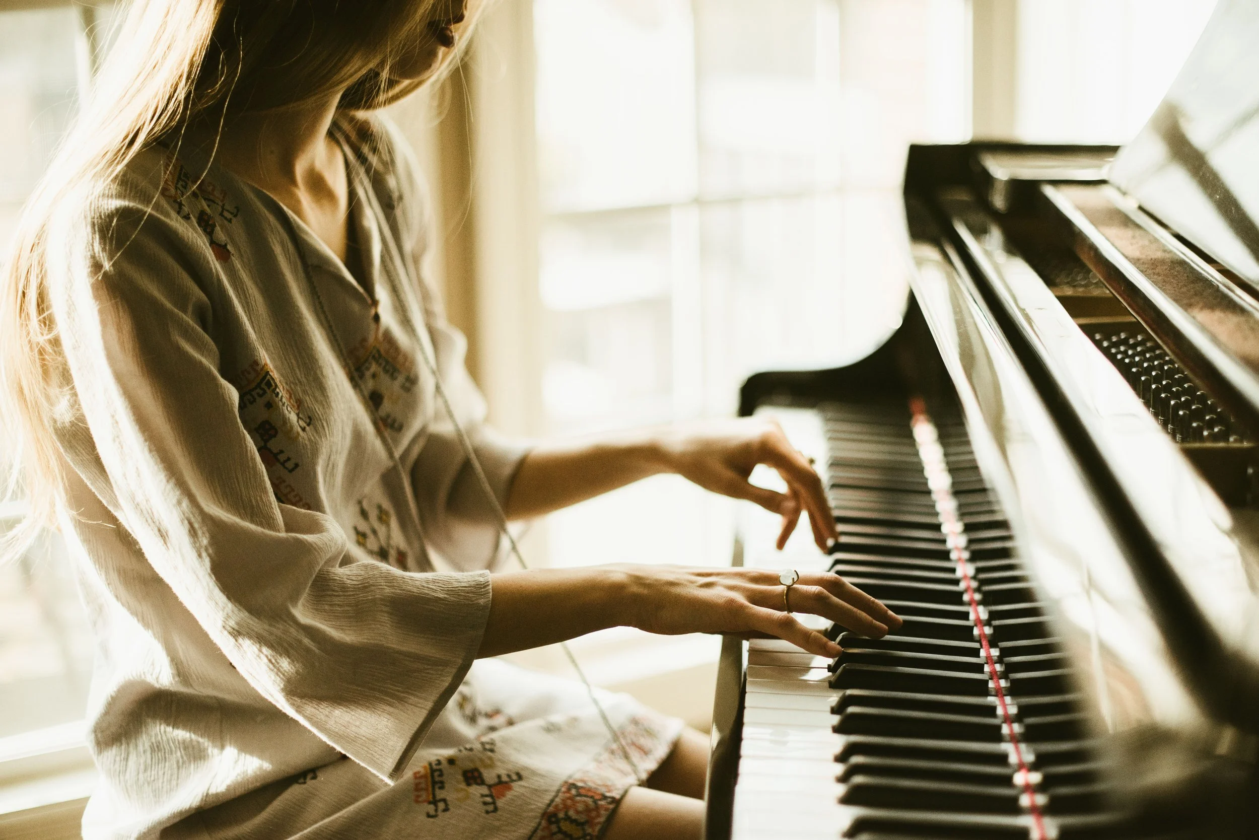 A woman with long blonde hair playing a grand piano indoors with sunlight streaming through the windows.