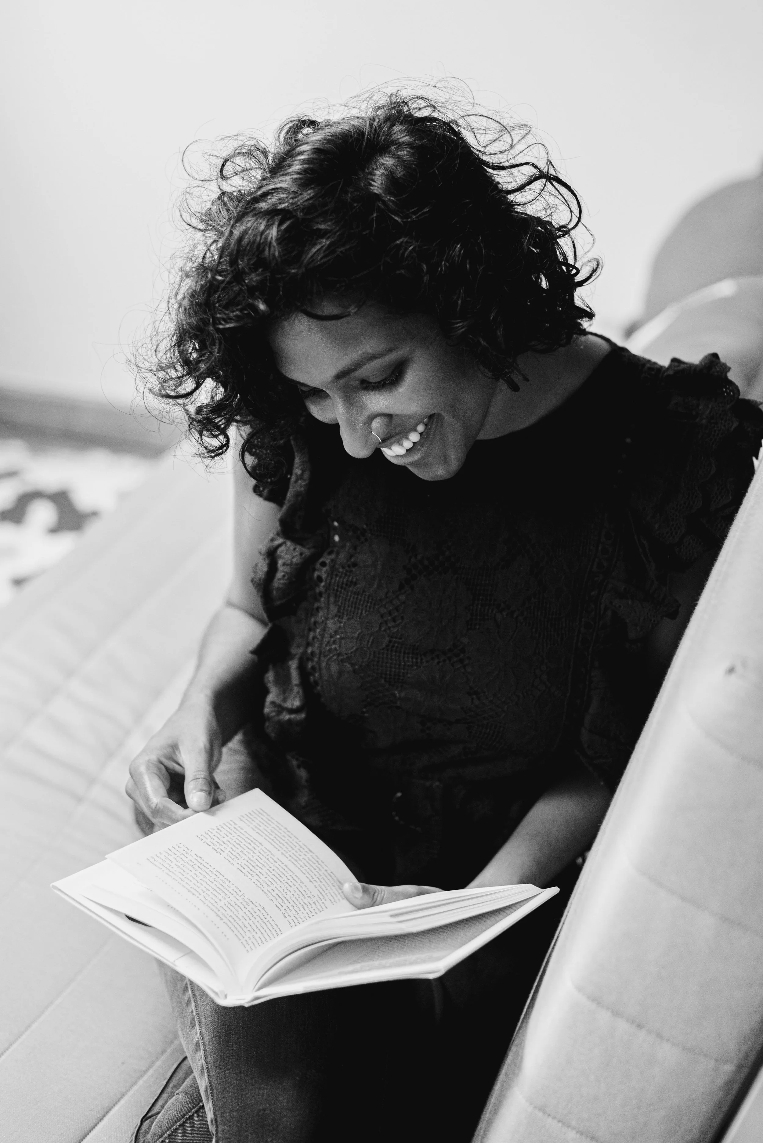 A woman with curly hair and a nose ring laughing while reading a book, sitting on a chair.