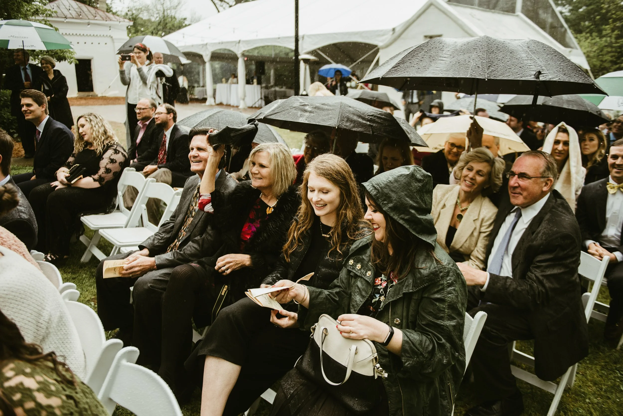 People sitting outdoors at a rainy event with umbrellas, some smiling, some taking pictures, in front of tents and white structures.
