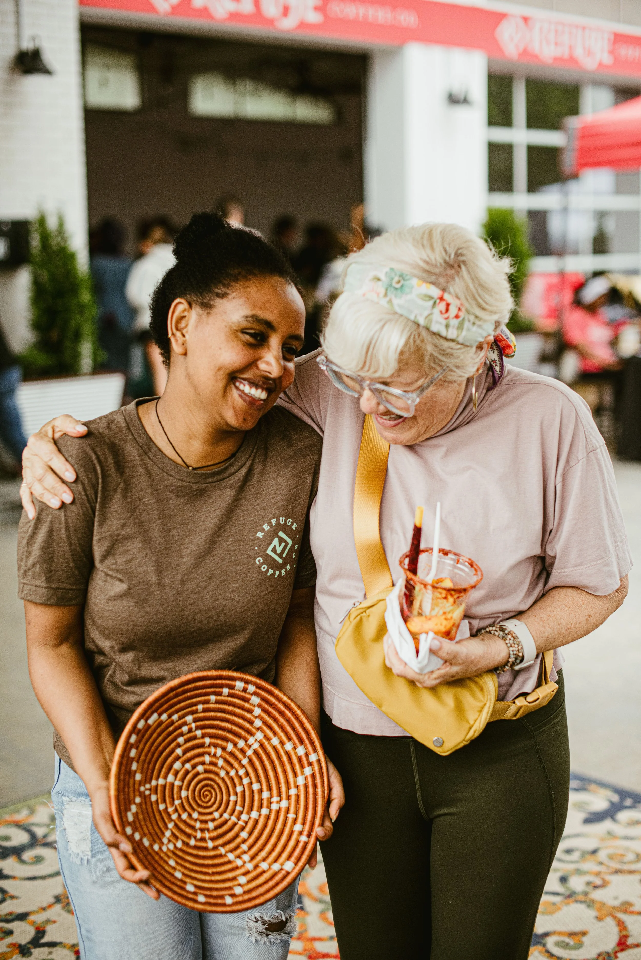 Two women smiling and hugging each other, one holding a bowl of pizza, in a lively indoor setting with people in the background.
