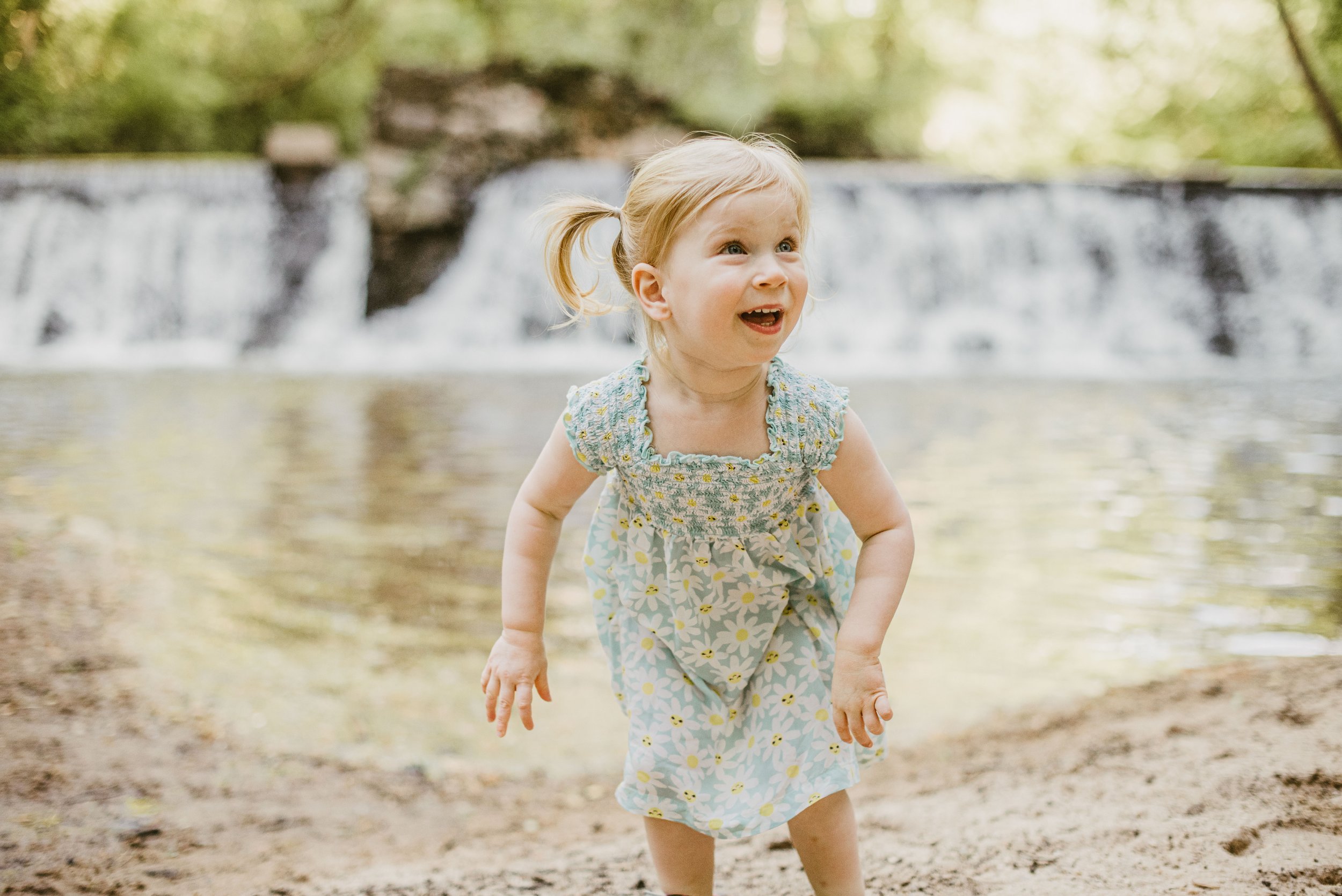 A young girl with blonde hair in a ponytail, wearing a floral dress, smiling and playing in a shallow creek near a small waterfall in a forest.