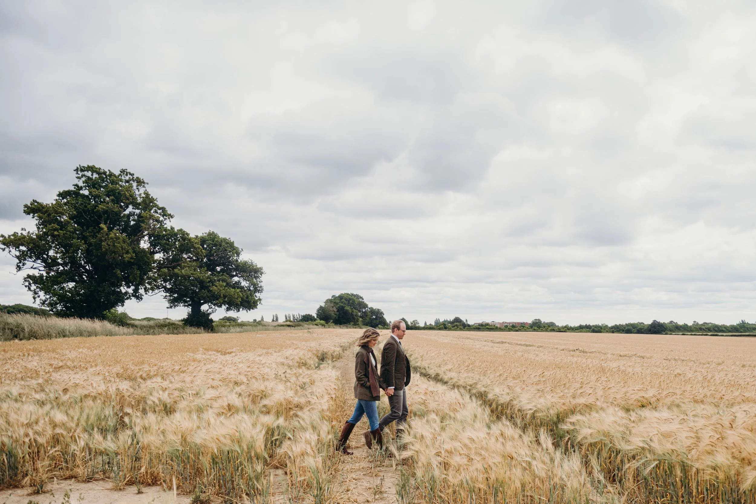 Two people walking through a field of wheat under cloudy skies, with large trees and a distant town in the background.