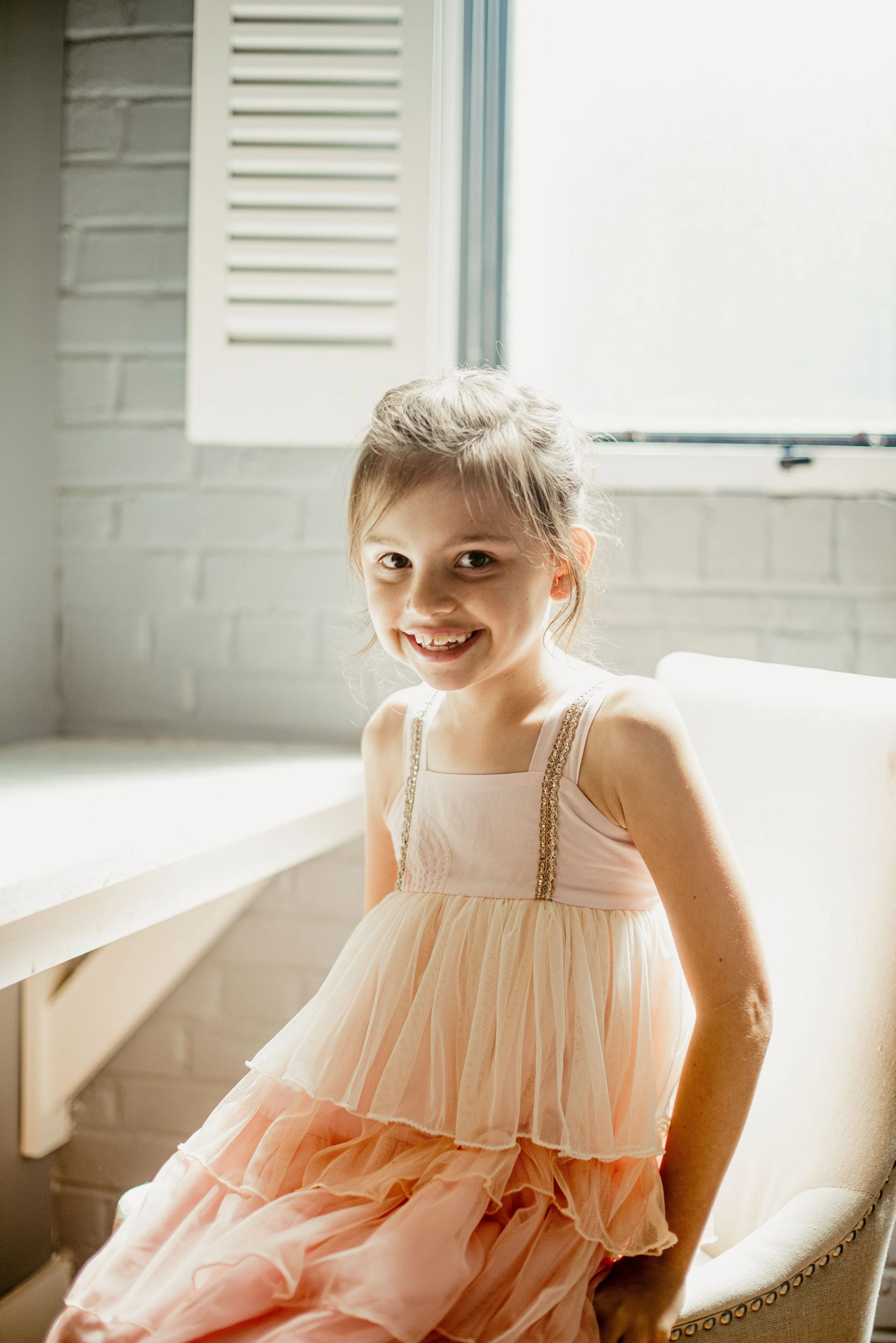 A young girl with light brown hair in a ponytail, wearing a light pink, sleeveless dress with a tiered skirt, standing inside near a window with sunlight streaming in, smiling at the camera.