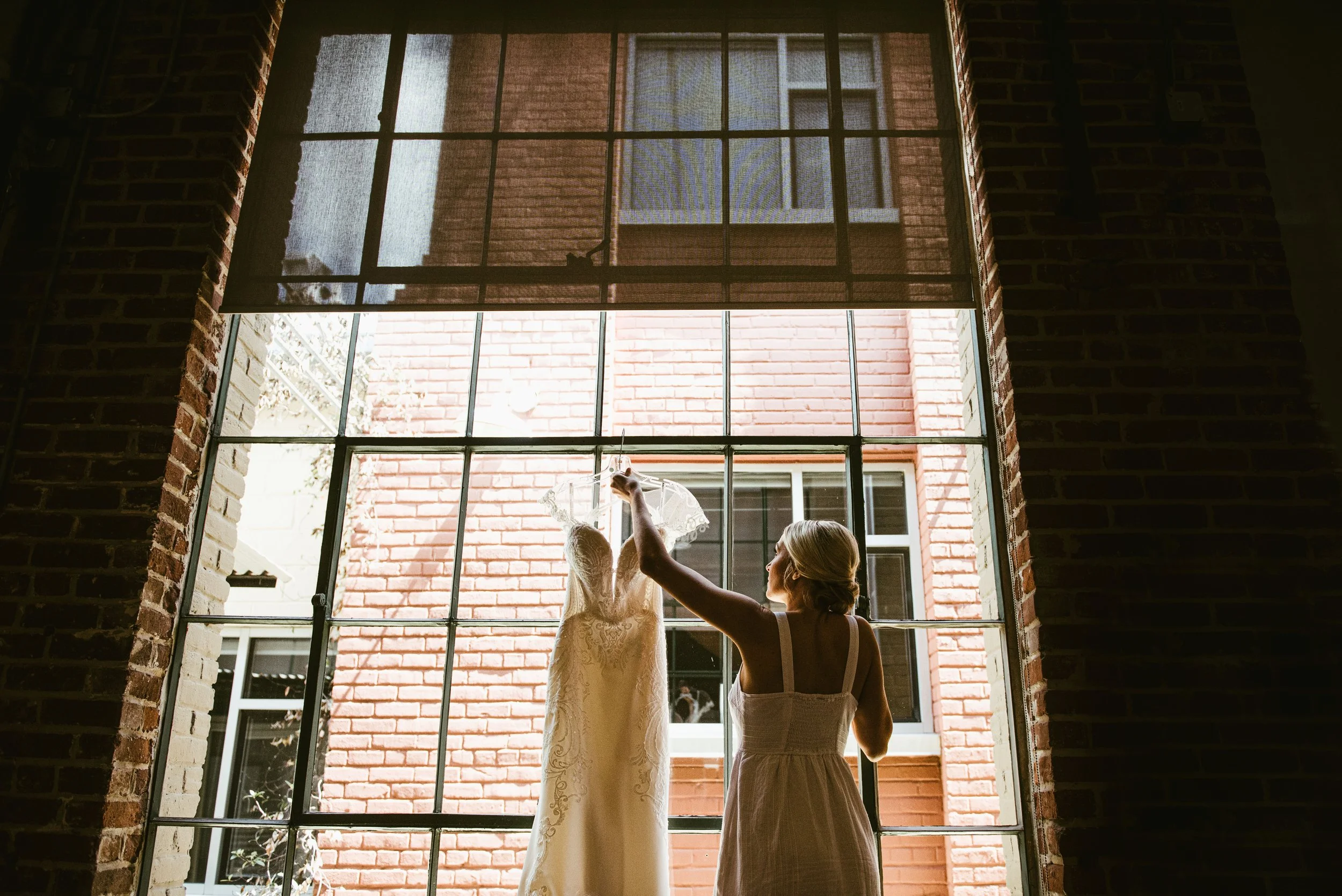 A woman and a girl are standing by a large window with a grid pattern. The woman is assisting the girl with a lace dress, possibly preparing for a special occasion. The scene is backlit by daylight, with brick walls surrounding the window.