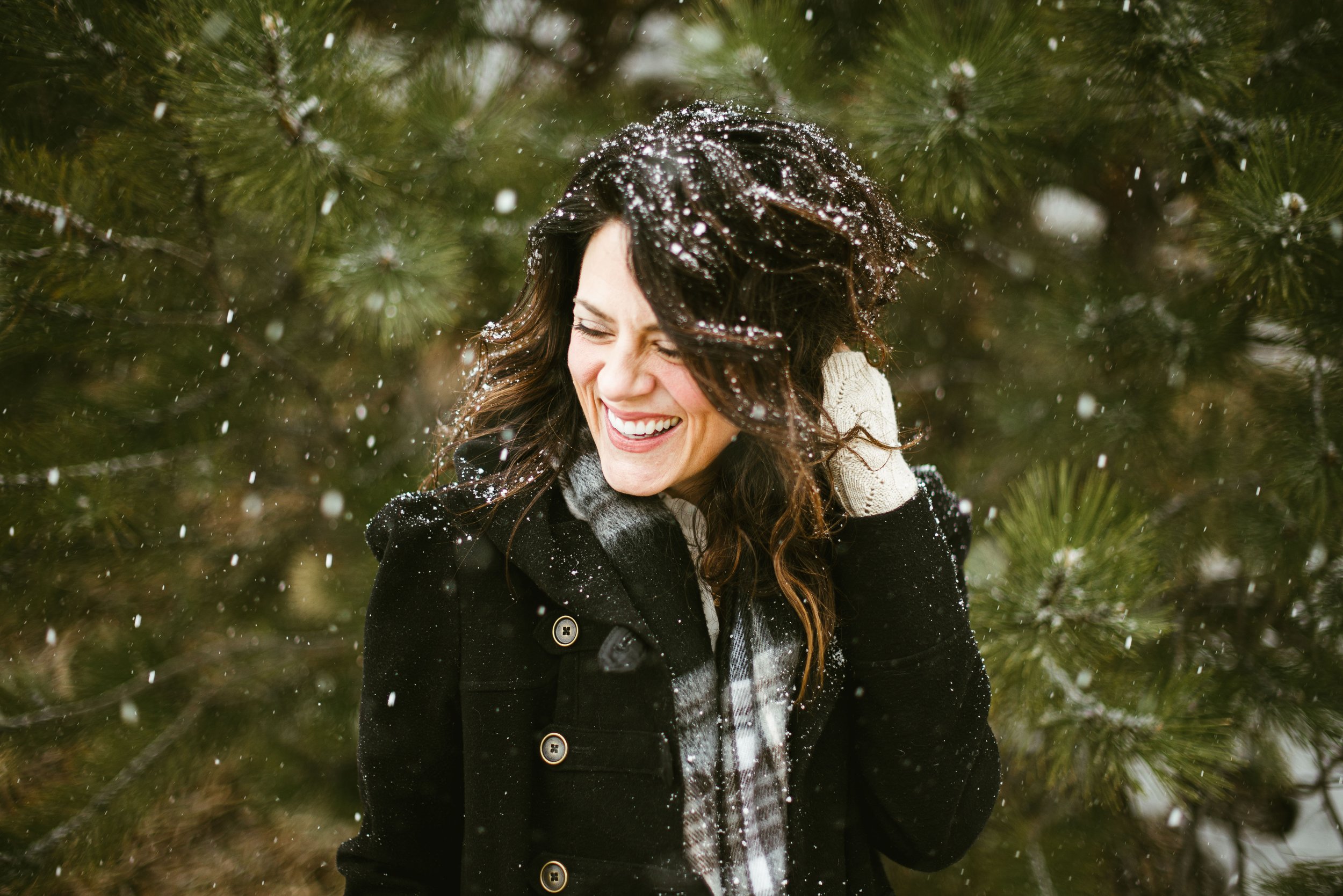 A woman with long dark hair, smiling, standing outdoors among green pine trees with snow falling around her.