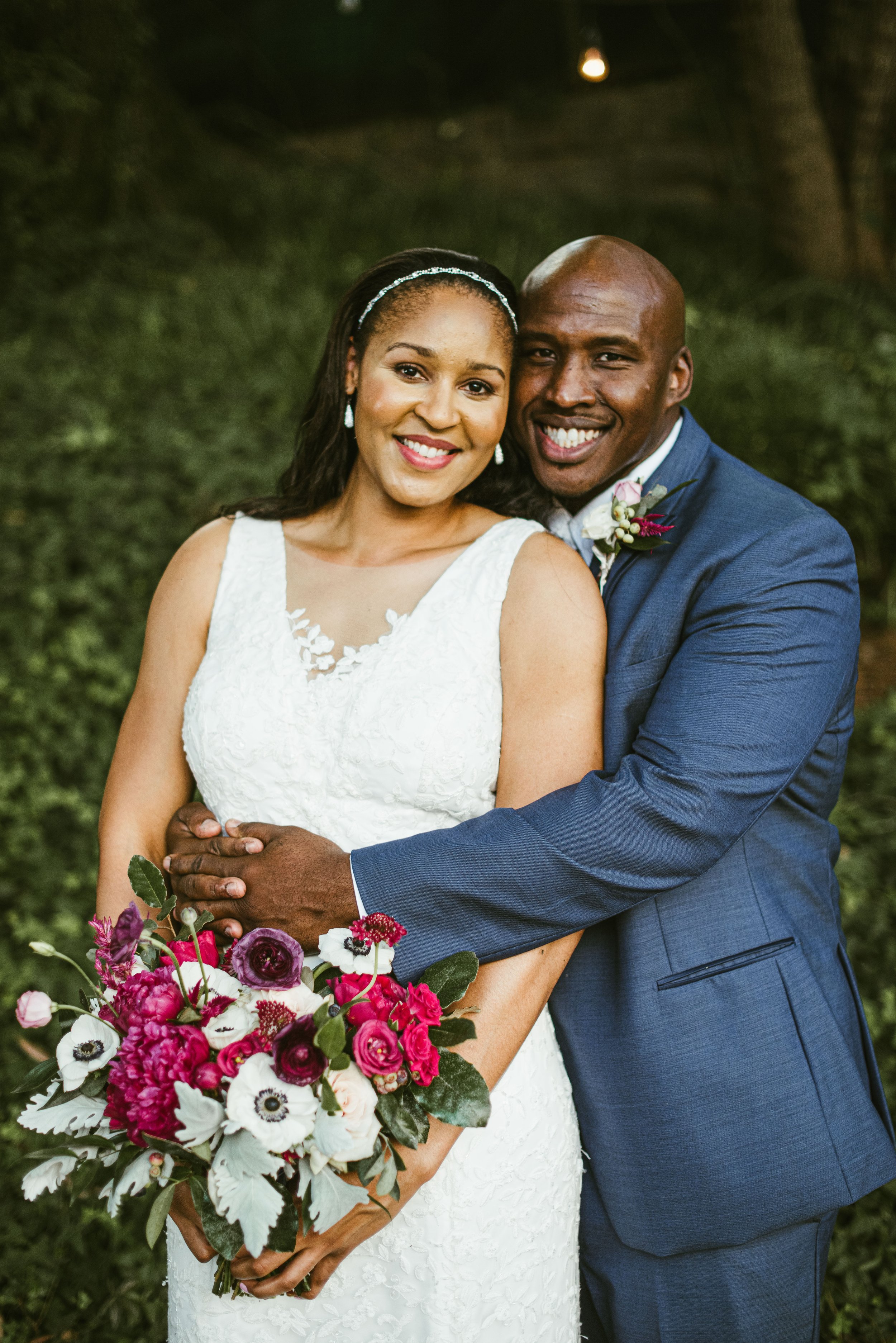 A smiling couple in wedding attire, standing outdoors with greenery in the background. The woman in a white sleeveless wedding dress holding a colorful bouquet, and the man in a blue suit with a boutonniere, hugging as they pose for a photo.