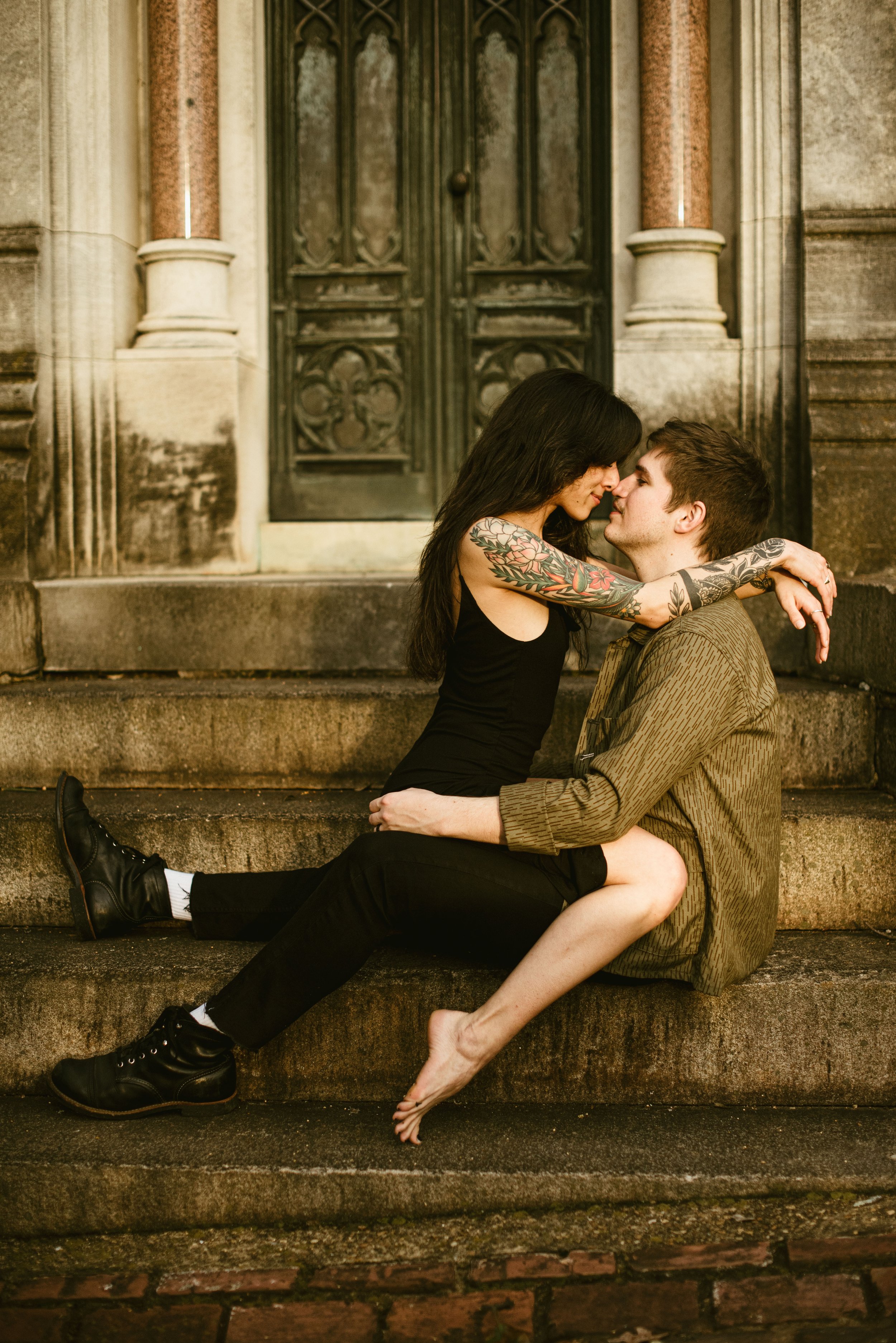 A couple sitting on stone steps in front of a large ornate wooden door, embracing and leaning towards each other, with a romantic vibe.
