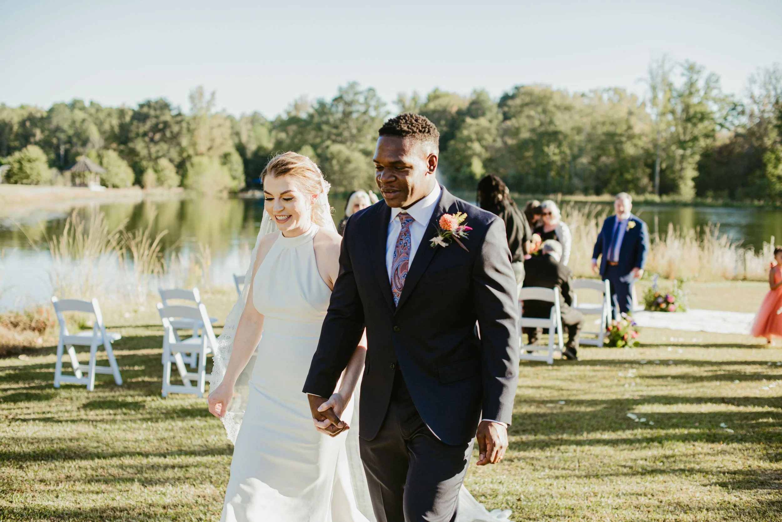 A bride and groom holding hands and walking outdoors at their wedding ceremony near a lake with trees in the background.