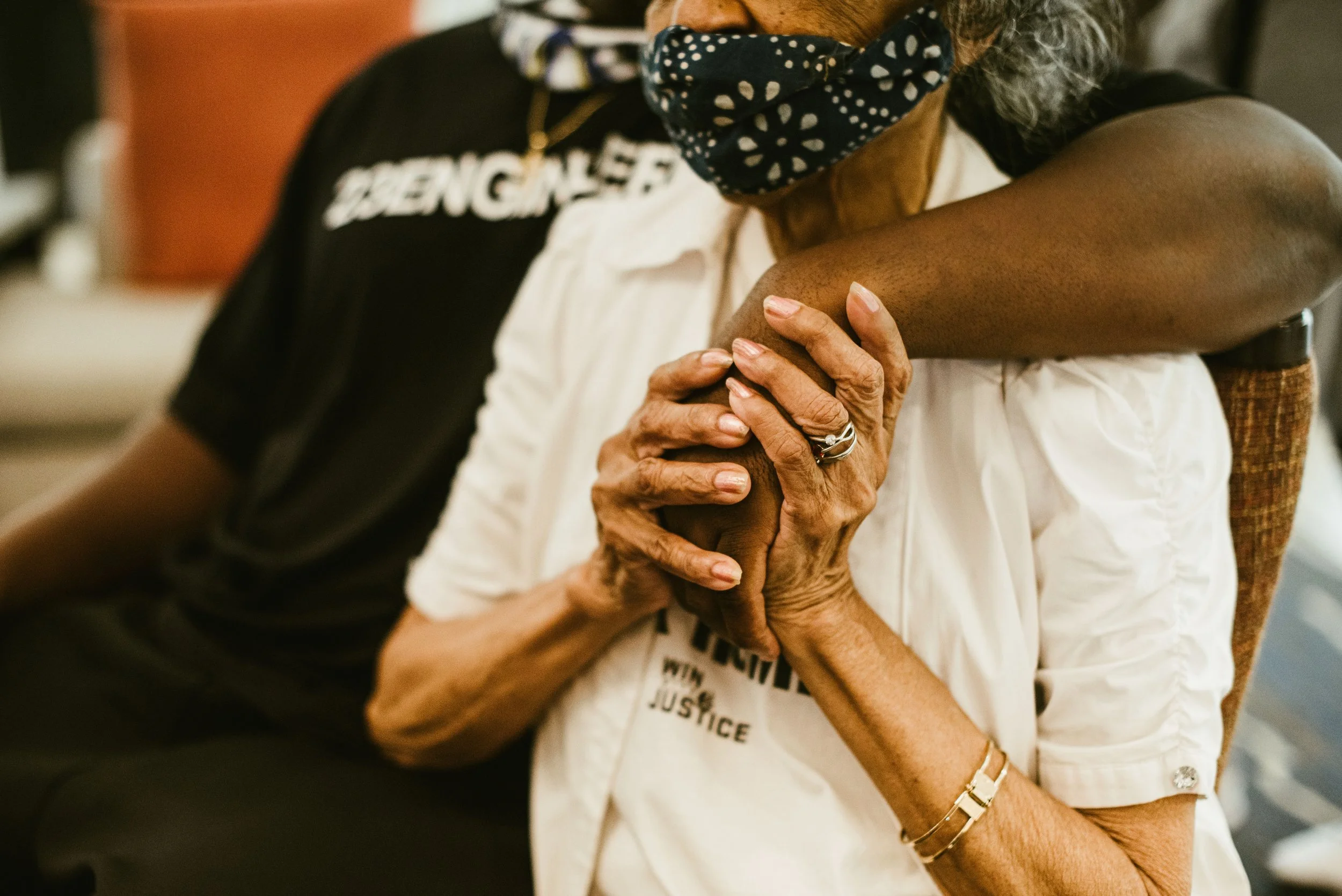 Close-up of an elderly woman's arm resting on her lap, with her hand gently holding her arm. A person behind her is placing a hand on her arm in a comforting gesture. The elderly woman wears a white shirt and jewelry; the person behind wears a black 