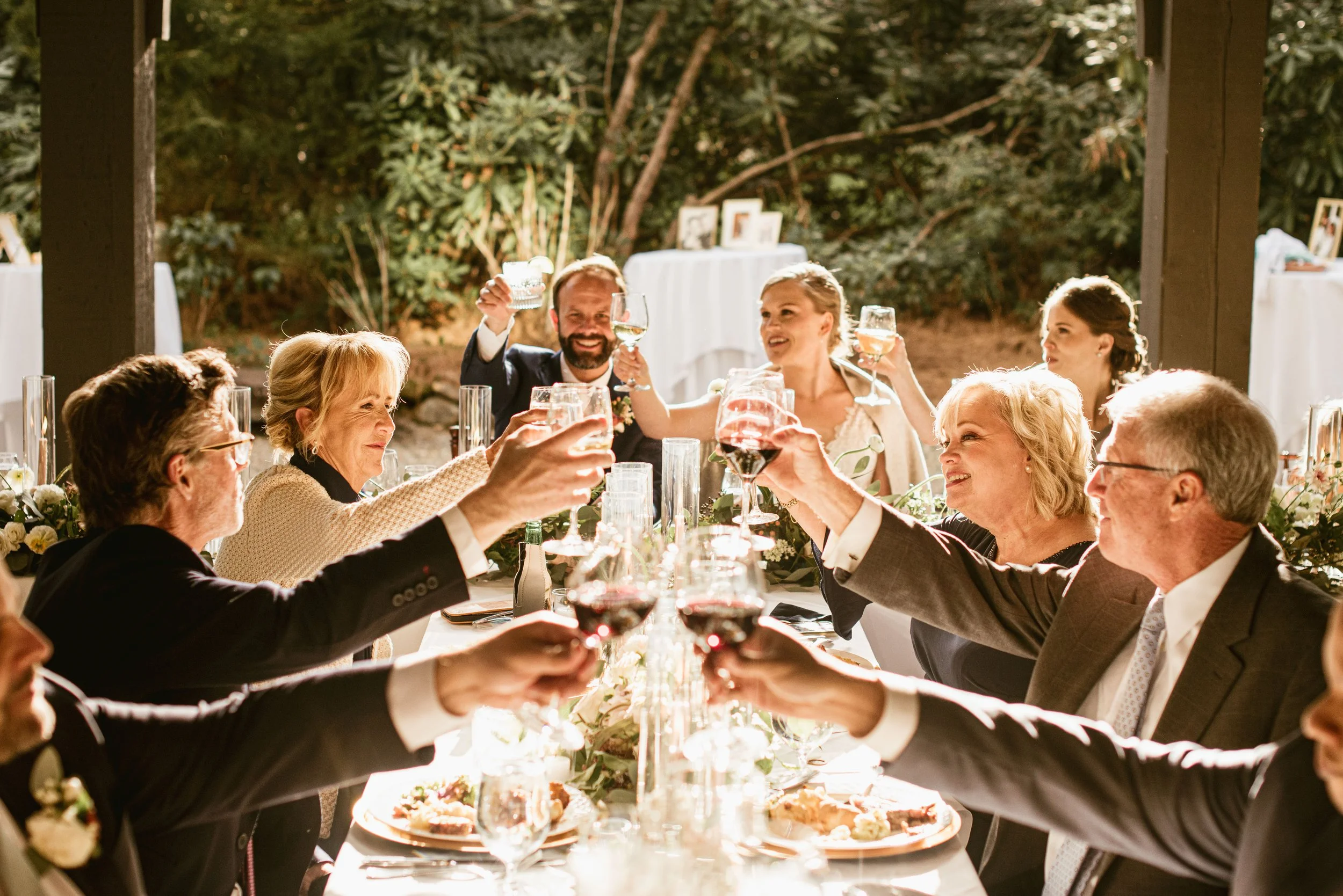 Group of people celebrating at a dinner party outdoors, raising glasses and toasting, with a lush green background and decorated dining table.