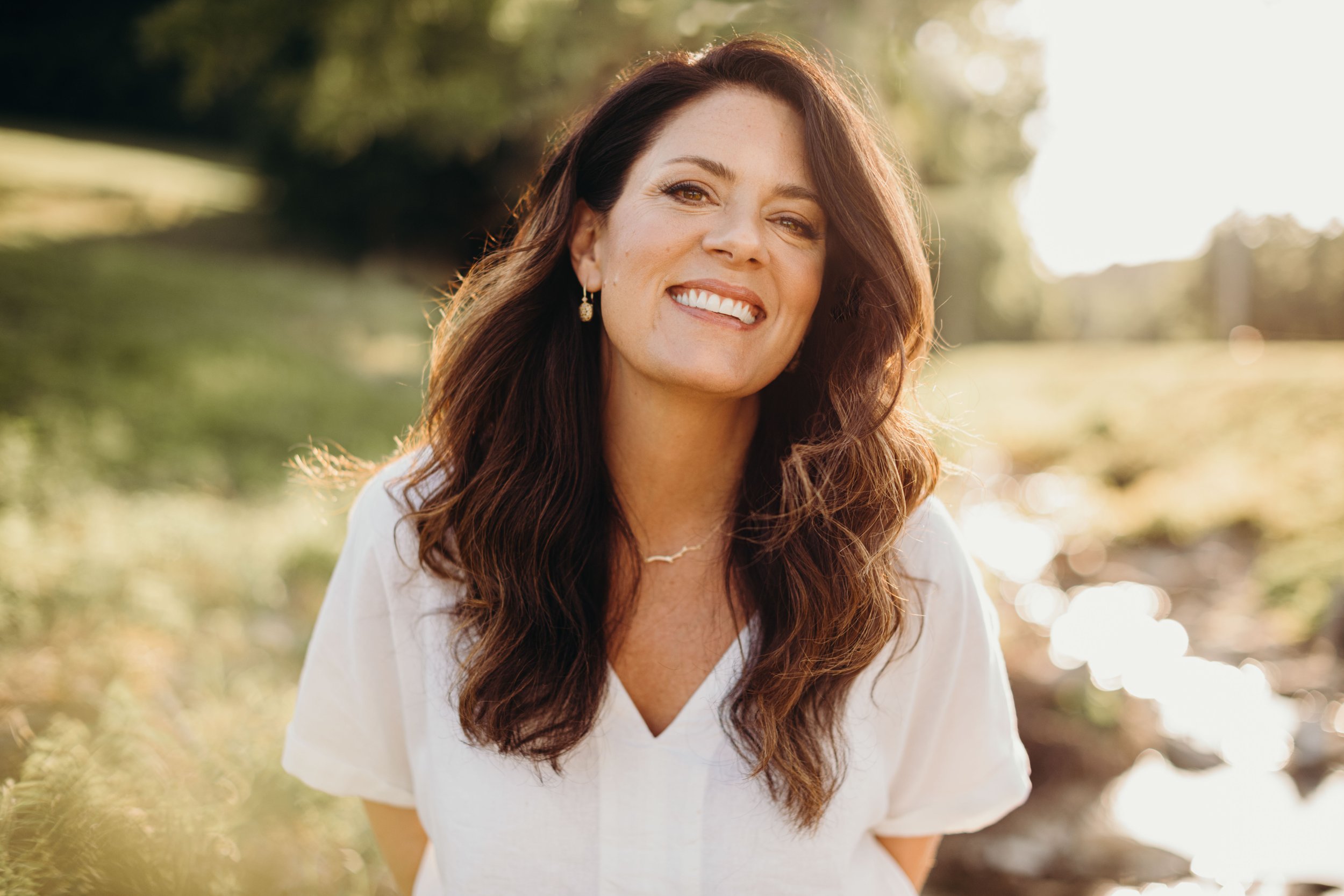 Close-up of a smiling woman with long, wavy brown hair, wearing earrings and a white top, outdoors with bright sunlight and blurred greenery in the background.