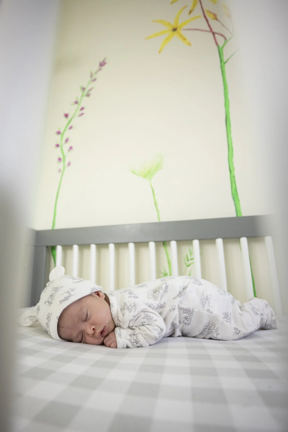A sleeping baby dressed in white lying on a bed with a gray and white checkered sheet, beneath a wall with colorful abstract floral artwork.