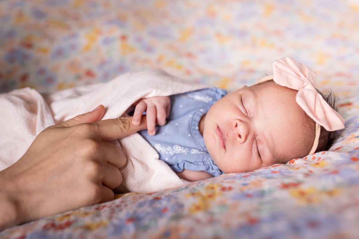 A sleeping baby girl with a pink bow headband, lying on a colorful blanket, being gently touched on the hand by an adult.