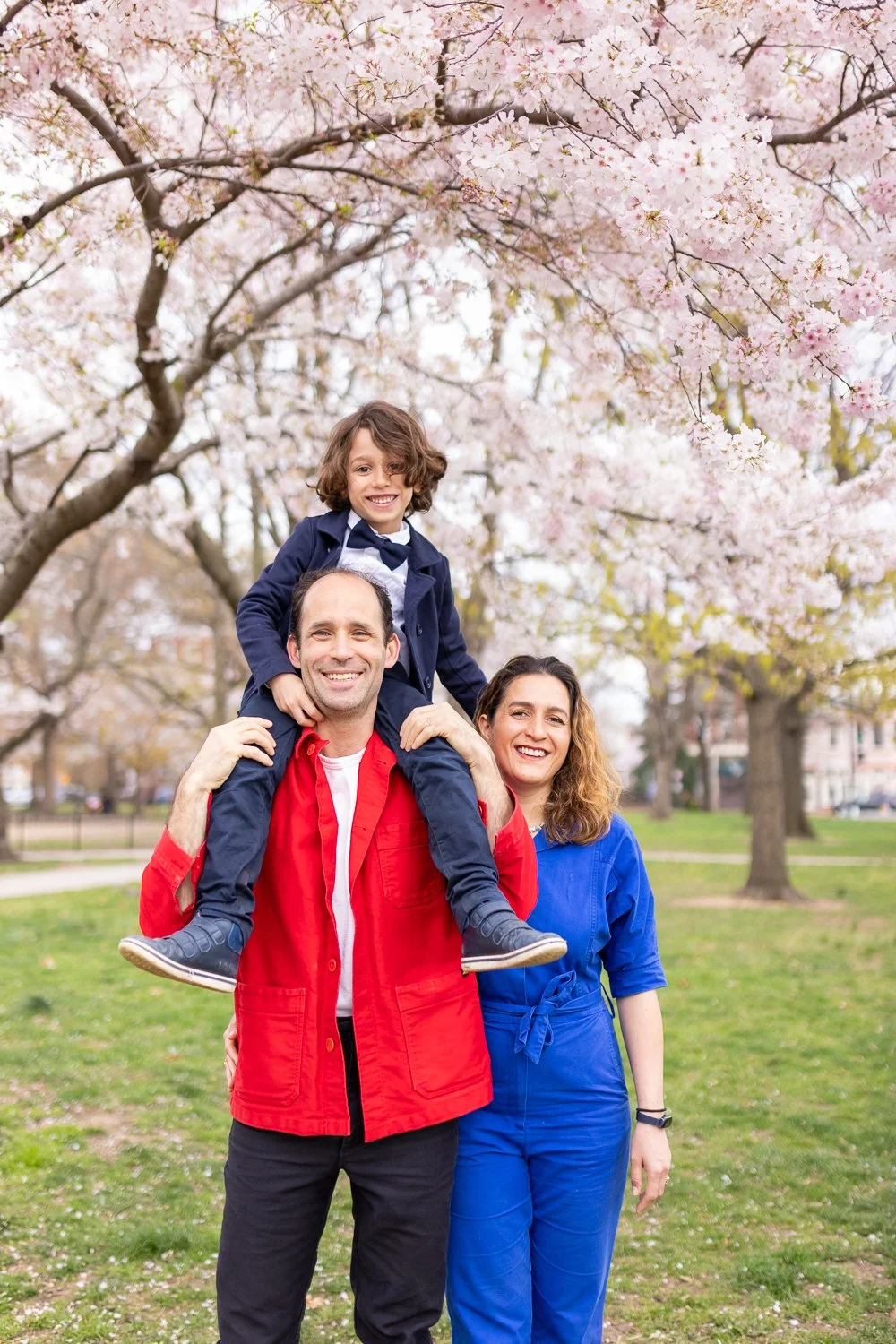 Happy family of three enjoying a day outdoors under blooming cherry blossom trees. The father is carrying the young boy on his shoulders, and the mother is standing beside them smiling.