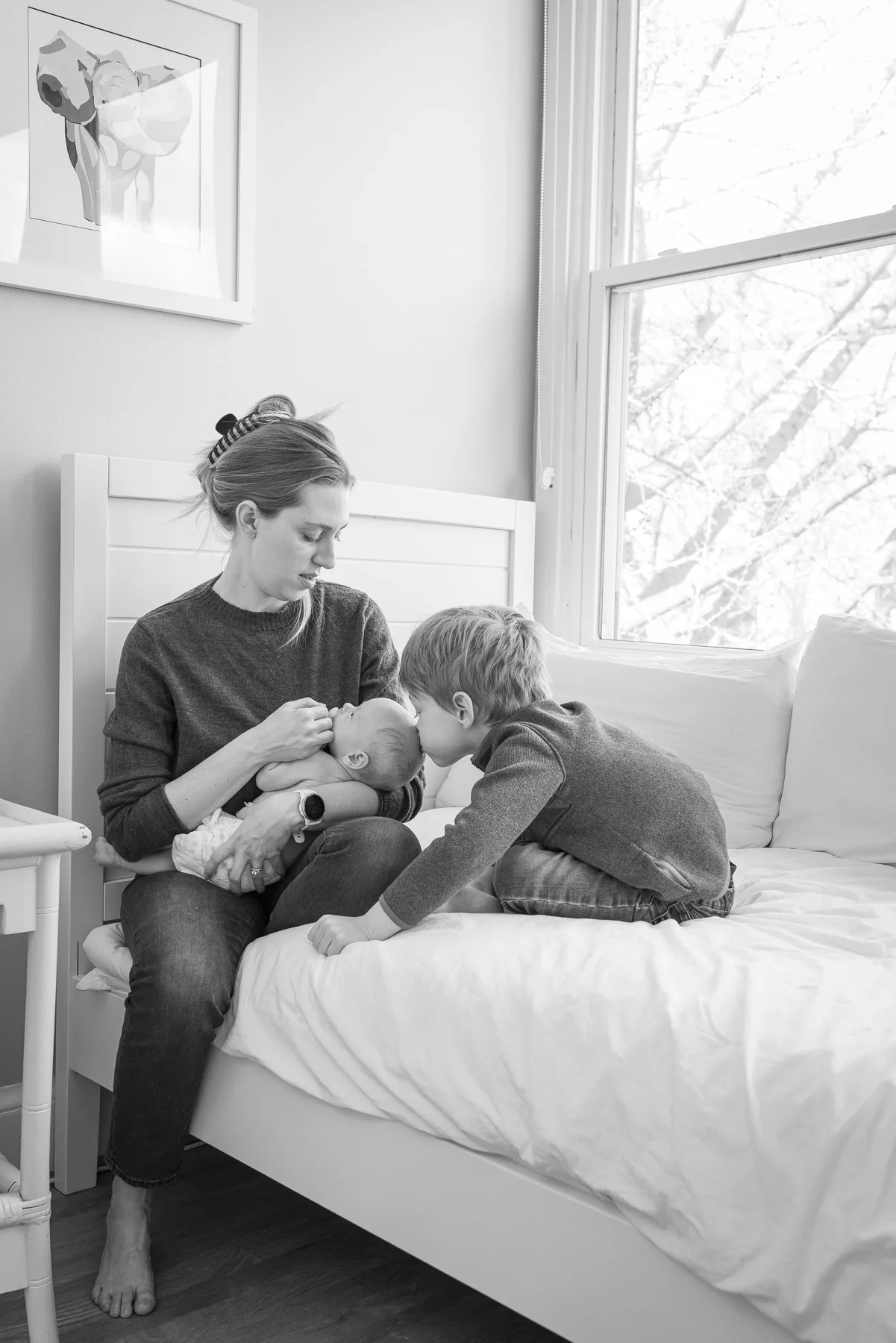Newborn lifestyle portrait on a bed with mom and older sibling kissing baby's head.