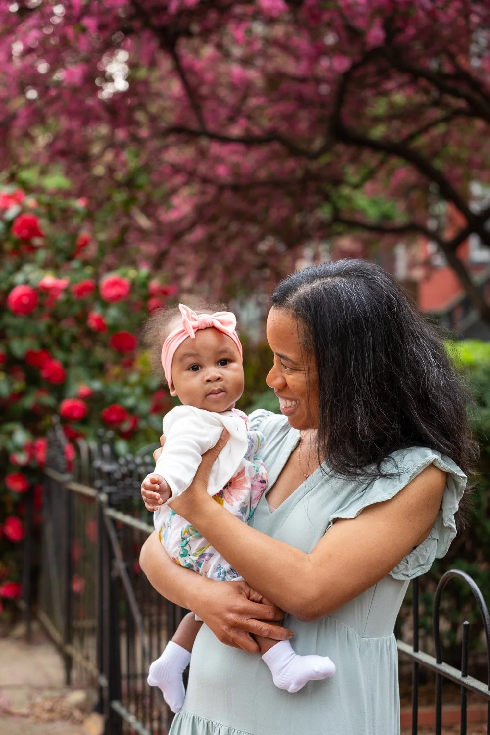 A woman holding a baby girl outdoors in front of a flowering pink tree and red roses, both smiling and enjoying a moment together.