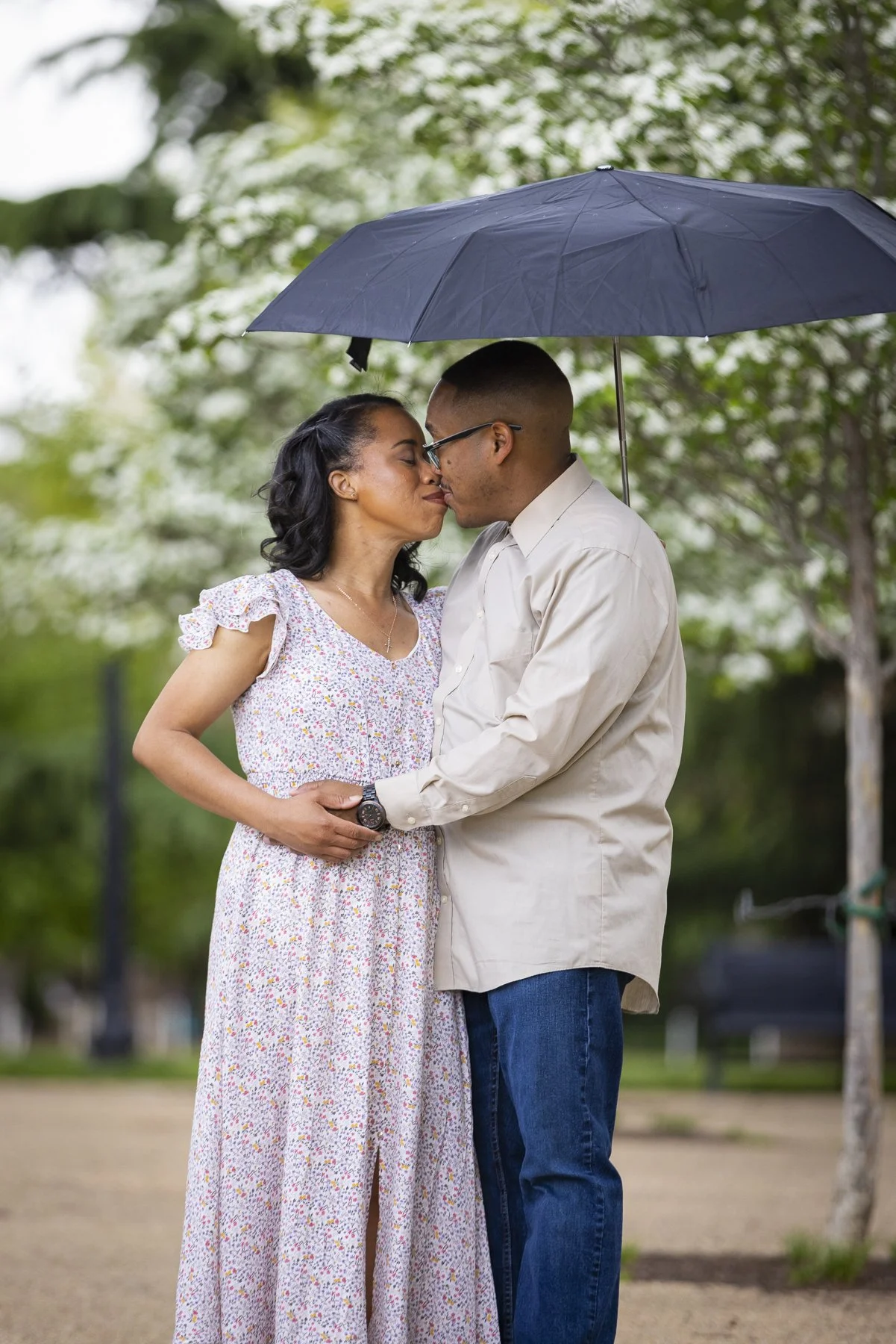 A couple sharing a kiss under a black umbrella in a park during rainy weather.