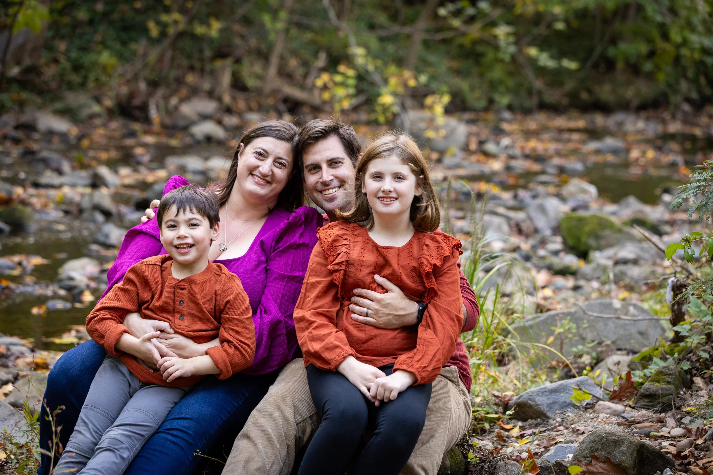 A family of four sitting outdoors by a creek in a forest during fall, all smiling and close together. The adults are a woman in a purple shirt and a man, with the woman and two children, dressed in fall clothing, sitting on rocks near the water.
