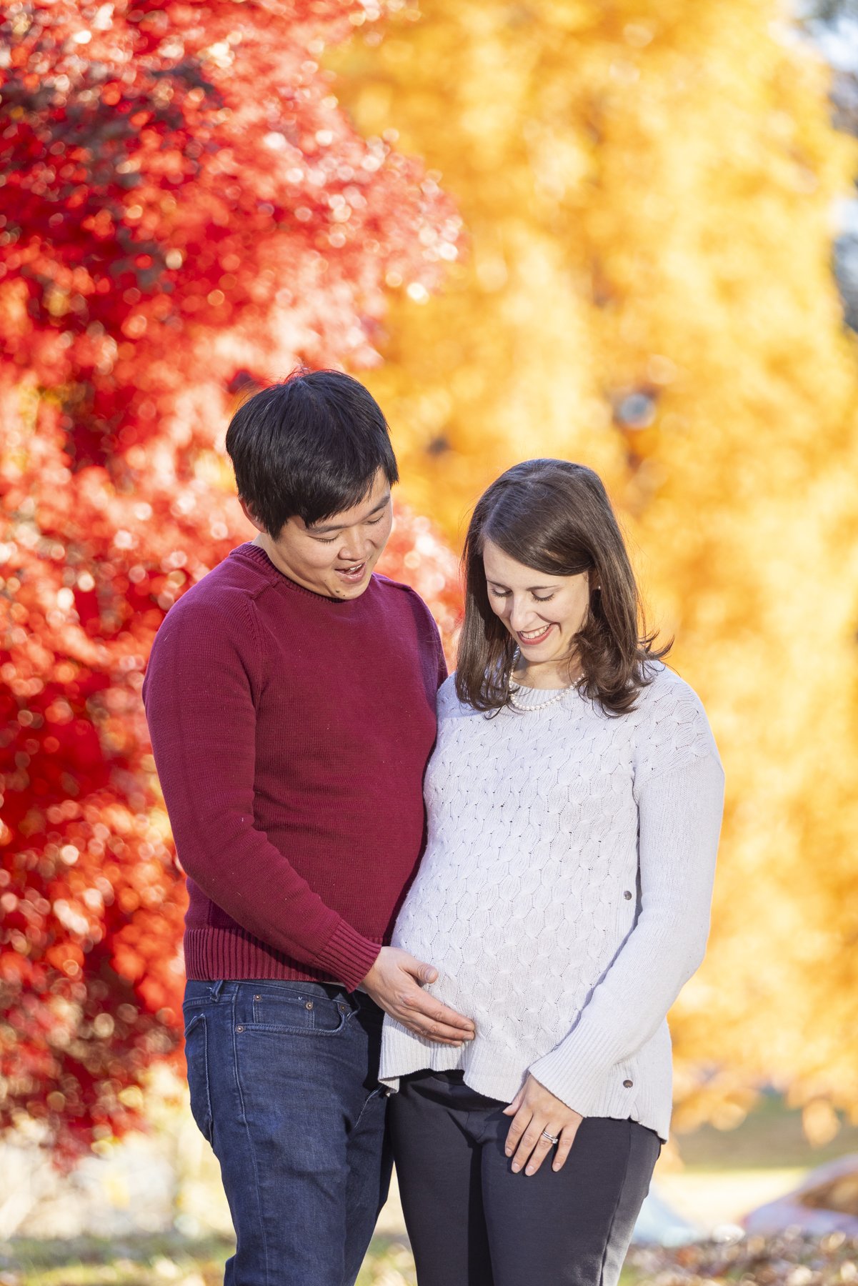 A couple, with the man gently touching the woman's pregnant belly, standing outdoors in fall with colorful autumn trees in the background.