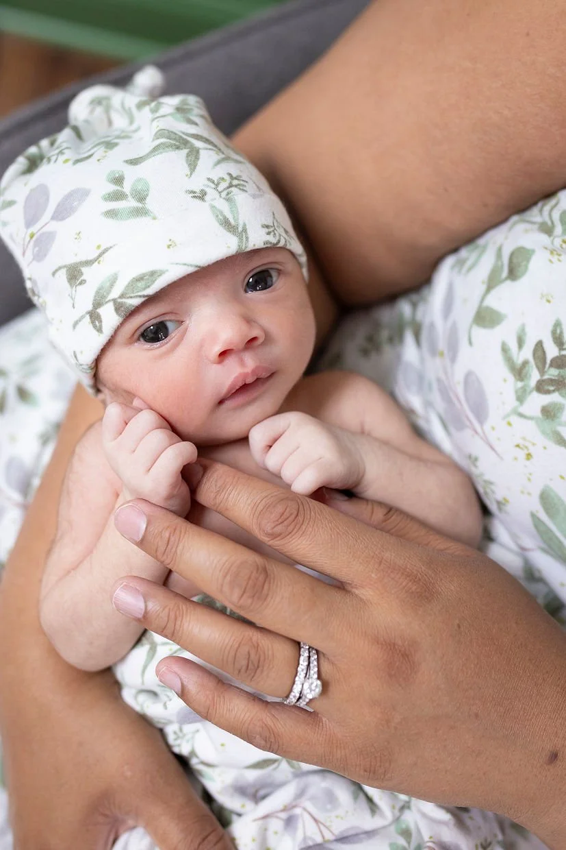 Close-up of a newborn baby lying on an adult's arm, wearing a white hat and outfit with green leaves, and looking at the camera.