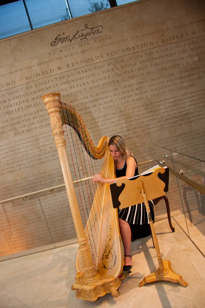 A woman playing a large wooden harp in front of a wall with inscriptions about the Donald W. Reynolds Foundation and other benefactors.