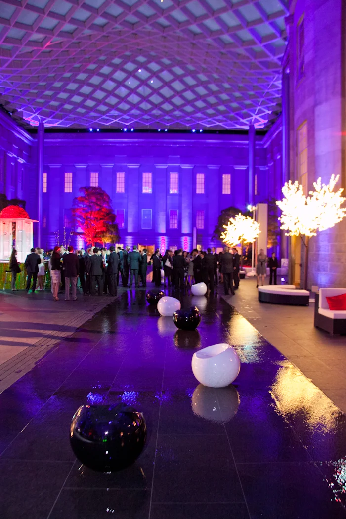 Indoor event space with purple and blue lighting, modern white and black decorative spherical objects, illuminated tree-like structures, and a crowd of people in formal attire.