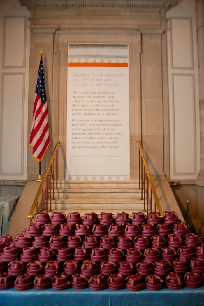 A display of multiple red graduation caps with tassels arranged on a table in front of the Corcoran Gallery of Art and College of Art and Design, with an American flag and an informational sign on a staircase in the background.