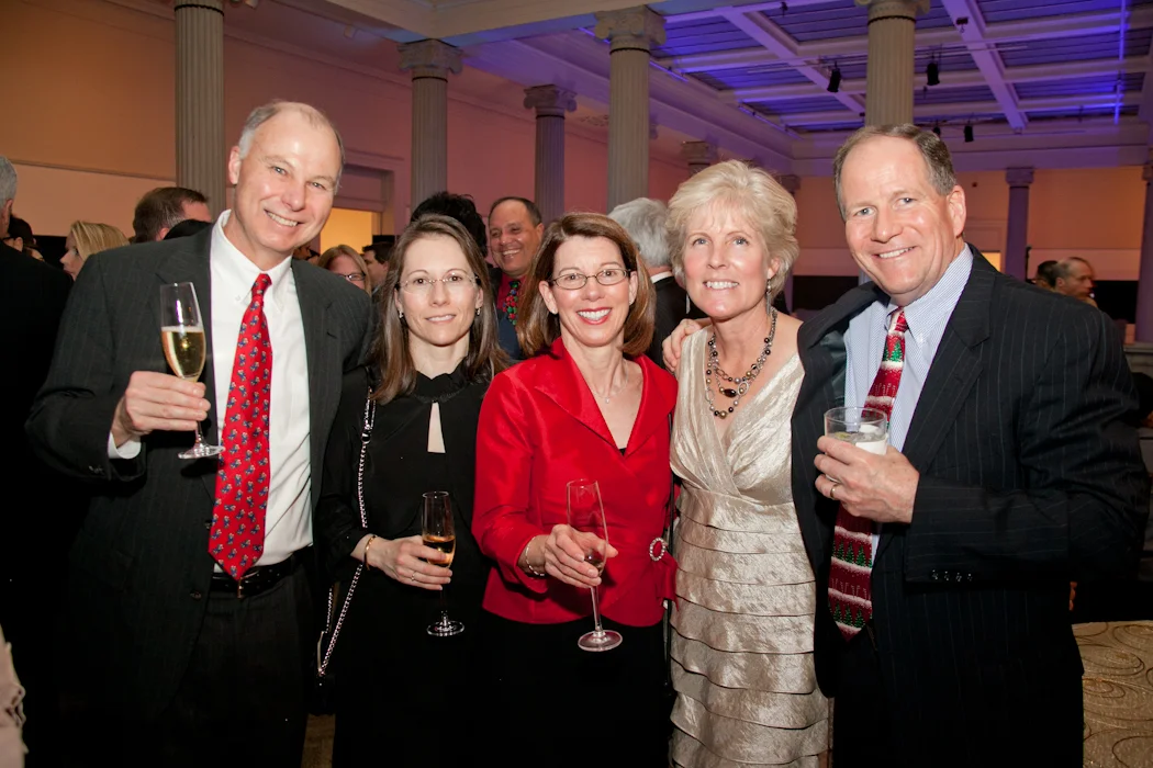 Group of five adults dressed in formal attire, smiling and holding drinks at a social event in a decorated venue with columns and purple lighting.