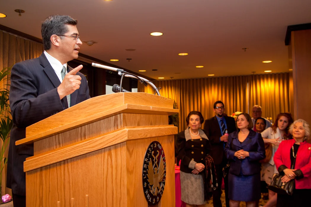 A man in a dark suit and glasses is speaking at a podium during an indoor event, with an audience of formally dressed women and men listening attentively in the background.