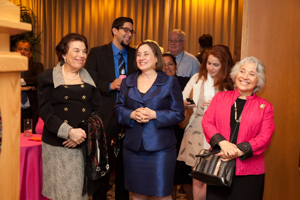Group of smiling people at a social event, including an older woman in a pink jacket, a woman in a blue suit, and a man in a suit with glasses, standing in a warmly lit room with curtains.
