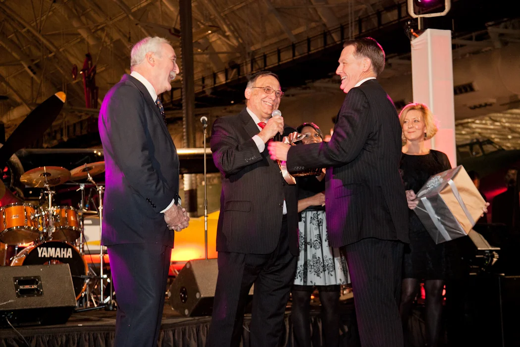 Group of five people in suits and dresses on a stage, with a drum set in the background, at an indoor event. One man is speaking into a microphone while another man hands him a gift, and others look on smiling.