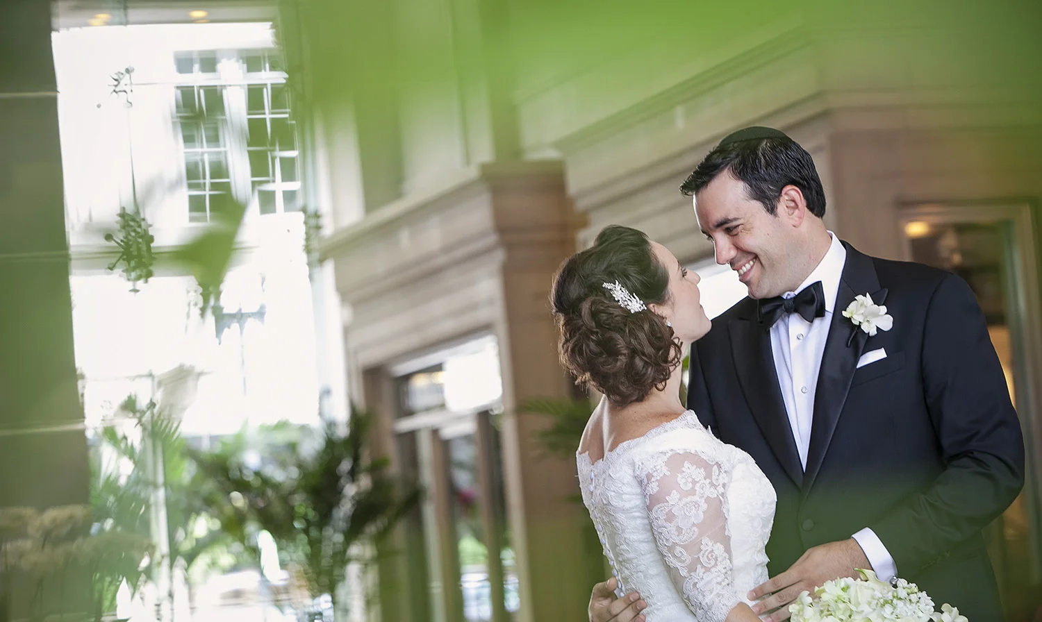 A bride and groom sharing a moment indoors at their wedding, smiling at each other, with the bride in a lace wedding dress and the groom in a black tuxedo with a bow tie.