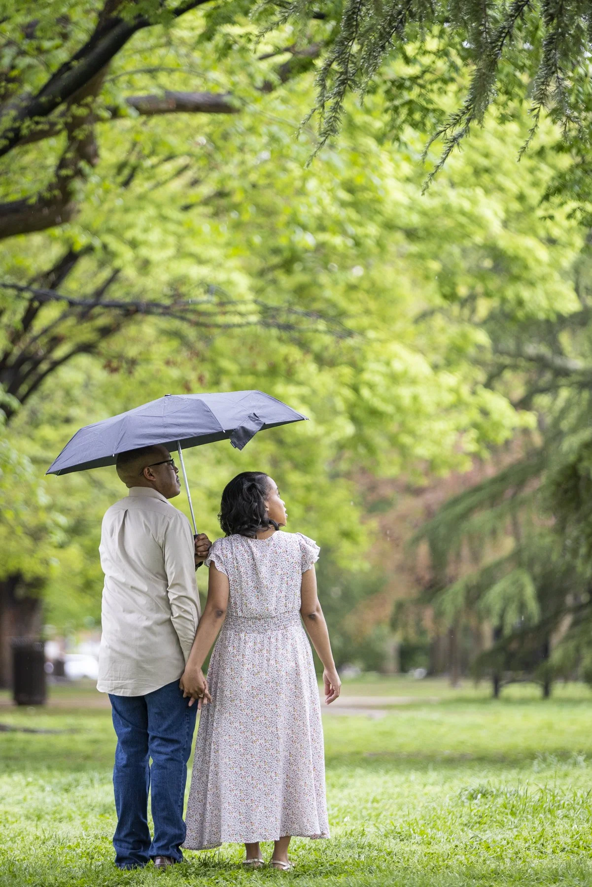 A man and a woman standing together in a park, holding hands; the man is holding an umbrella over both of them, and they are surrounded by lush green trees.