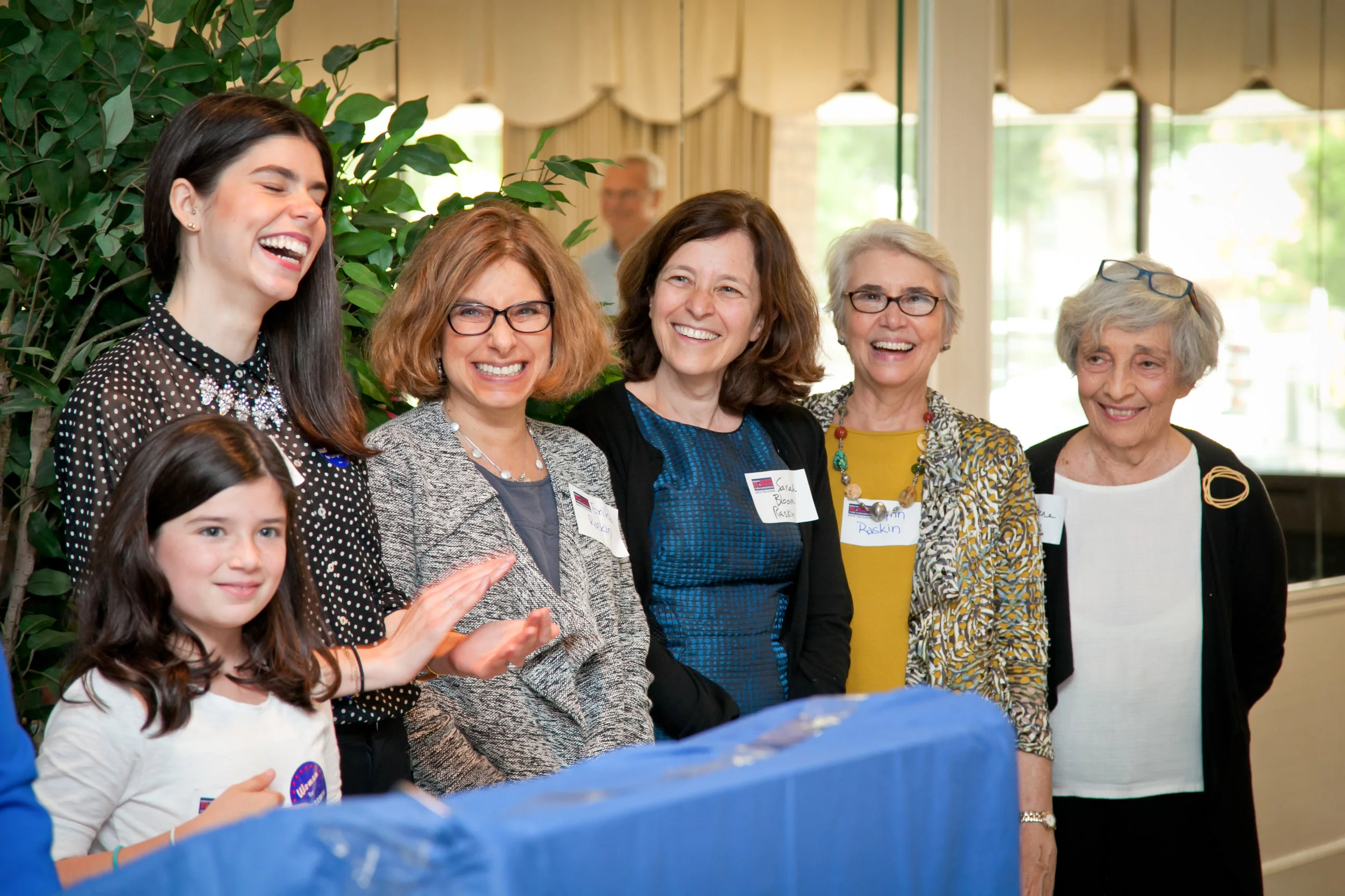 A group of women and a young girl are gathered together, smiling and laughing at an indoor congressional campaign event with large windows and green plants in the background.
