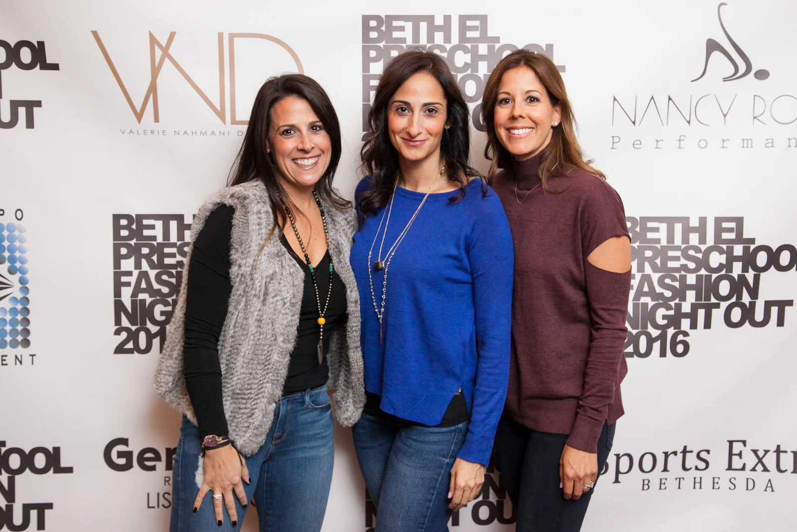 Three women standing together at a fashion event in front of a white backdrop with logos and text, smiling at the camera.