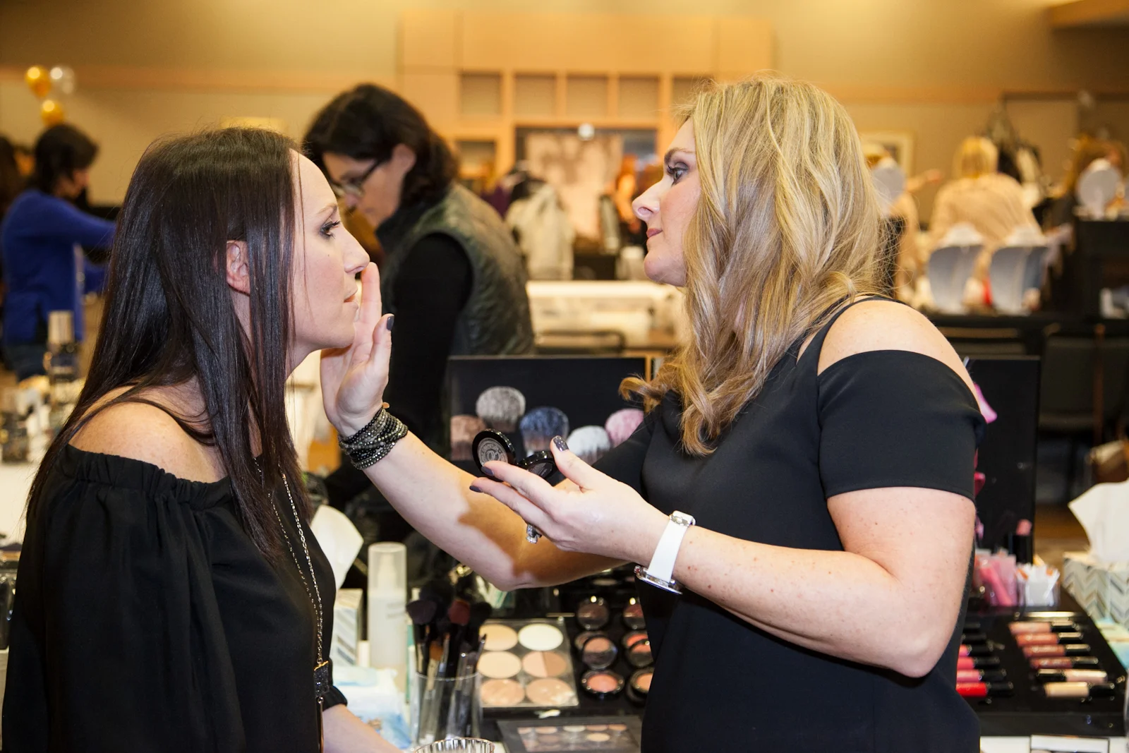 A woman applies makeup to another woman at a makeup booth during an event, with various makeup products visible on the table.